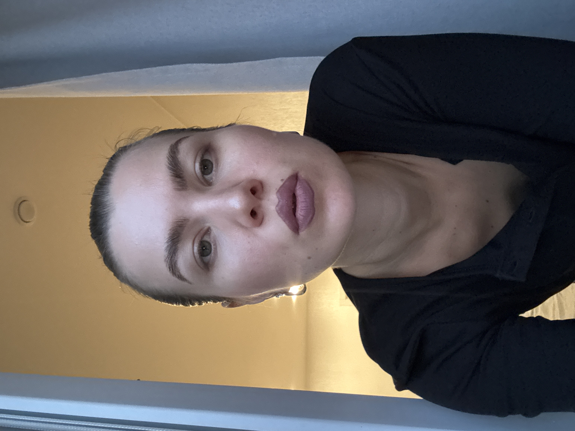 An overhead, medium shot captures a woman in a dark, long-sleeved shirt. She is looking directly at the camera with her lips pursed. The woman has full cheeks, visible pores, and a light dusting of freckles. Her eyebrows are dark and defined, and she is wearing a single, teardrop-shaped earring in her left ear. The lighting in the scene is warm and soft, casting a glow on her face from the left. In the background, a pale yellow wall is visible, along with a white curtain on the right side and a light blue vertical panel on the left. The ceiling has a circular light fixture. The scene appears to be indoors.