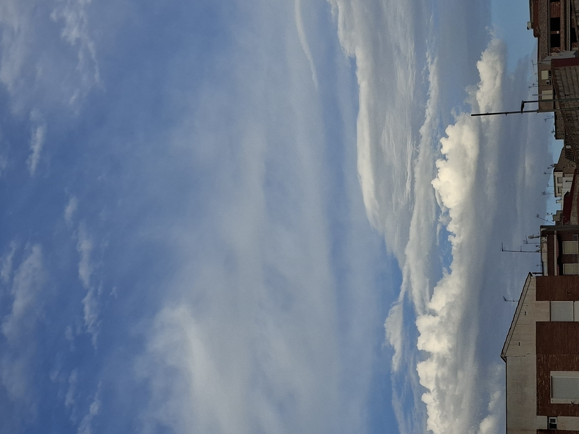 The image captures a sky over the city of Bullas, Spain, featuring a dynamic interplay of clouds against a blue sky. The lower portion of the frame reveals the rooftops of buildings, suggesting an urban or semi-urban environment.
The sky is dominated by varying cloud formations. High in the sky, wispy cirrus clouds stretch across the blue expanse, interspersed with patches of lighter blue. Further down, thicker, cumulus-like clouds are present, some appearing bright white and illuminated by sunlight, while others are tinged with grey, indicating depth and potentially varying atmospheric conditions. Some of these clouds have a layered, stratiform appearance.
The buildings in the foreground are varied in construction. The most prominent building on the left has a light-colored upper section and a brick-patterned lower facade. It features windows or shutters. To the right, other buildings are visible, including one with a darker roof and windows, and another with a more complex rooftop structure featuring antennae and satellite dishes. A tall, dark pole, possibly a utility pole or antenna mast, stands vertically in the middle-ground, piercing the skyline. The overall impression is of a typical daytime scene in a Spanish city, with architectural elements defining the earthly horizon against a vast sky.