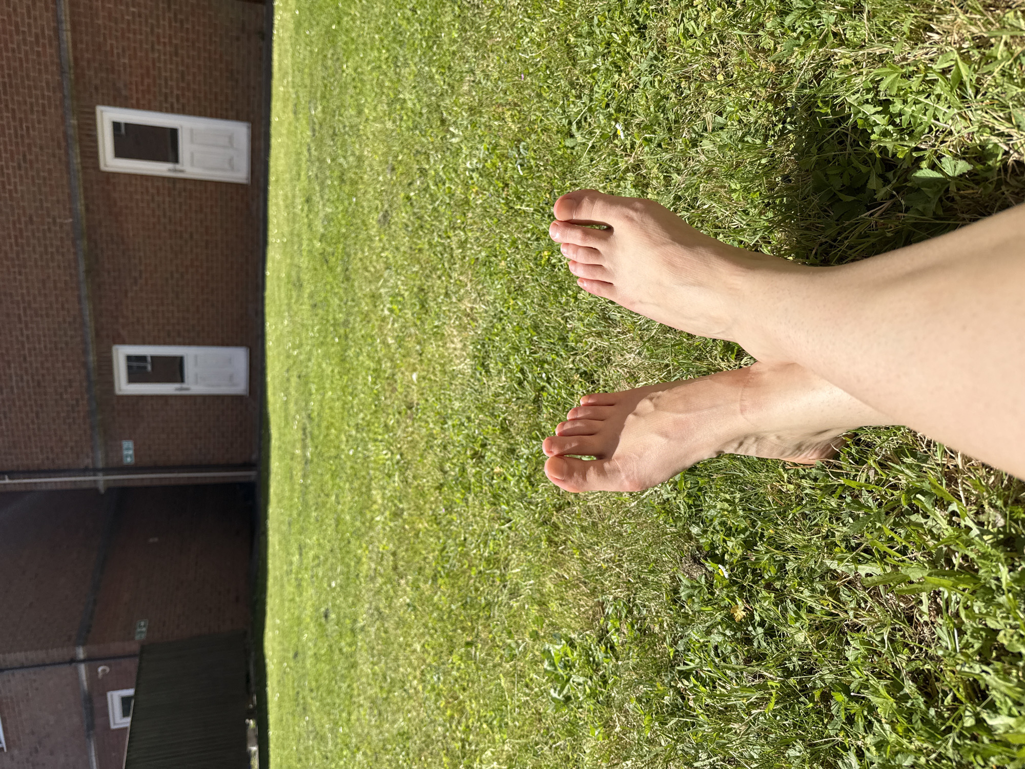 The image shows two bare feet crossed on a patch of green grass in an outdoor setting. The background features a brick building with two white windows. The location is Great Yarmouth, United Kingdom.