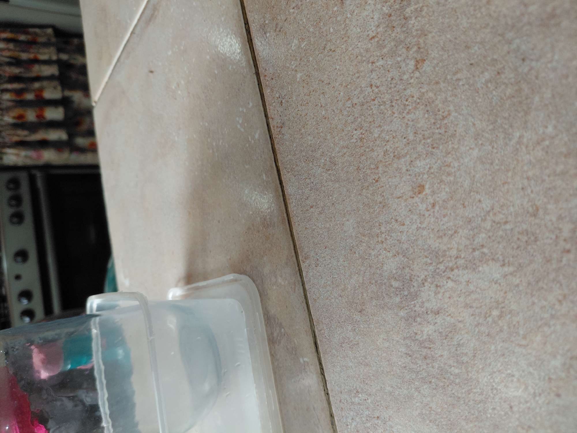 The image captures a close-up, rotated view of an indoor setting, likely a kitchen or utility area. In the foreground, two transparent plastic containers are visible. The container on the left holds a mix of pink and blue objects or liquids. The container on the right appears to be empty or contains clear liquid. The containers rest on a light-colored, speckled tiled surface, possibly a countertop or floor, given the angle.
A prominent grout line runs diagonally across the image, separating two sections of similarly textured, beige or light brown tiles. The tile to the right of the grout line appears slightly darker and more uniformly speckled. In the blurry background on the left, an appliance with circular knobs (possibly a stove or oven) and a patterned object, perhaps fabric or a stack of items, can be vaguely discerned. No people are visible, and no specific activity is taking place beyond the static arrangement of the objects. There is no visible text. The lighting suggests an indoor environment, but the time of day is not determinable.