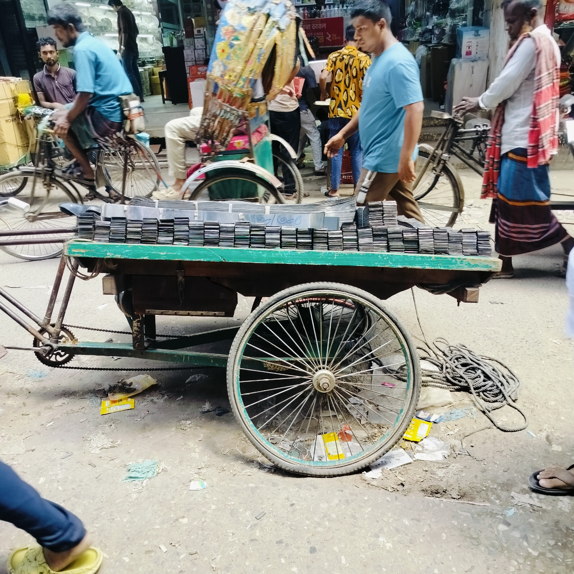 The image depicts a bustling street scene in Dhaka, Bangladesh. A three-wheeled cart, loaded with stacks of what appear to be aluminum sheets or compact discs, is the central focus. The cart has a green wooden frame and metal spoked wheels. Nearby, a coiled rope lies on the ground.
Several people are present in the background, including a man in a blue shirt walking past the cart, two men on bicycles, and another man wearing a patterned shawl, also walking. Other vehicles, including cycle rickshaws, are visible. The street is littered with debris. The setting suggests a commercial area, with shops visible in the background.