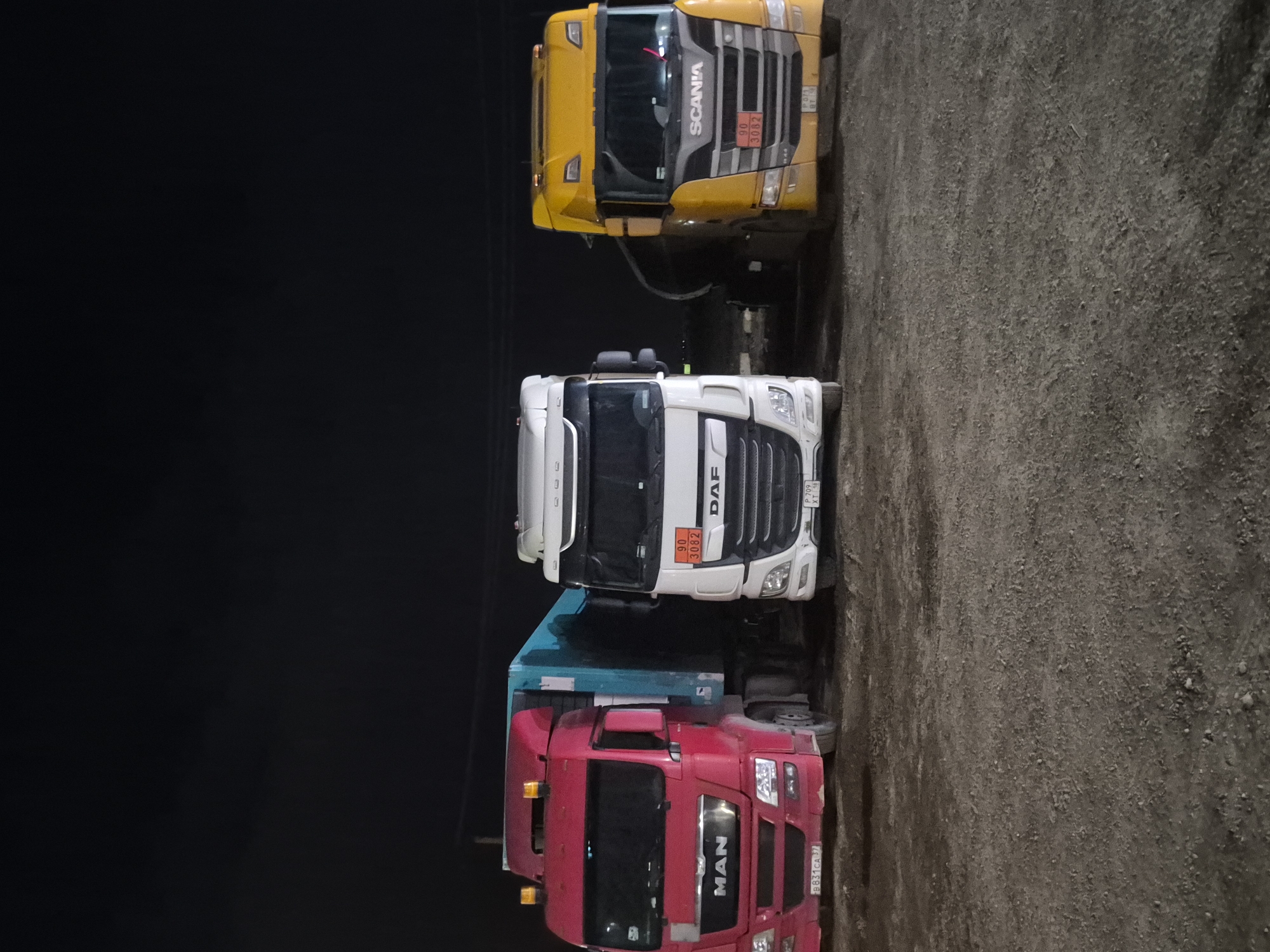 The image captures three large semi-trailer trucks parked side-by-side on a gravel or dirt surface at night in Mozhary, Russia. The sky is completely dark, indicating late evening or nighttime conditions. No people are visible, and there is no apparent activity taking place.
From left to right (as the image is rotated), the trucks are:
1. A bright yellow Scania truck. Its grille clearly displays "SCANIA". A hazard identification panel below the grille shows "90" on top and "3082" on the bottom, indicating a highly flammable liquid. A partial license plate "P01 XT 48" is visible.
2. A white DAF truck, positioned in the center. The "DAF" logo is prominent on its grille. Similar to the Scania, it also has a hazard identification panel with "90" over "3082". Its license plate reads "P 709 XT 46".
3. A red MAN truck, with its "MAN" logo displayed on the grille. This truck appears to be a dump truck or a truck with a blue container body attached behind the cabin, though the container is mostly obscured by darkness and the other truck. Its license plate is "B 831 CA 37".
The ground is rough, consisting of small stones and dirt. The trucks are parked closely together. The overall impression is of a resting or storage area for heavy vehicles.