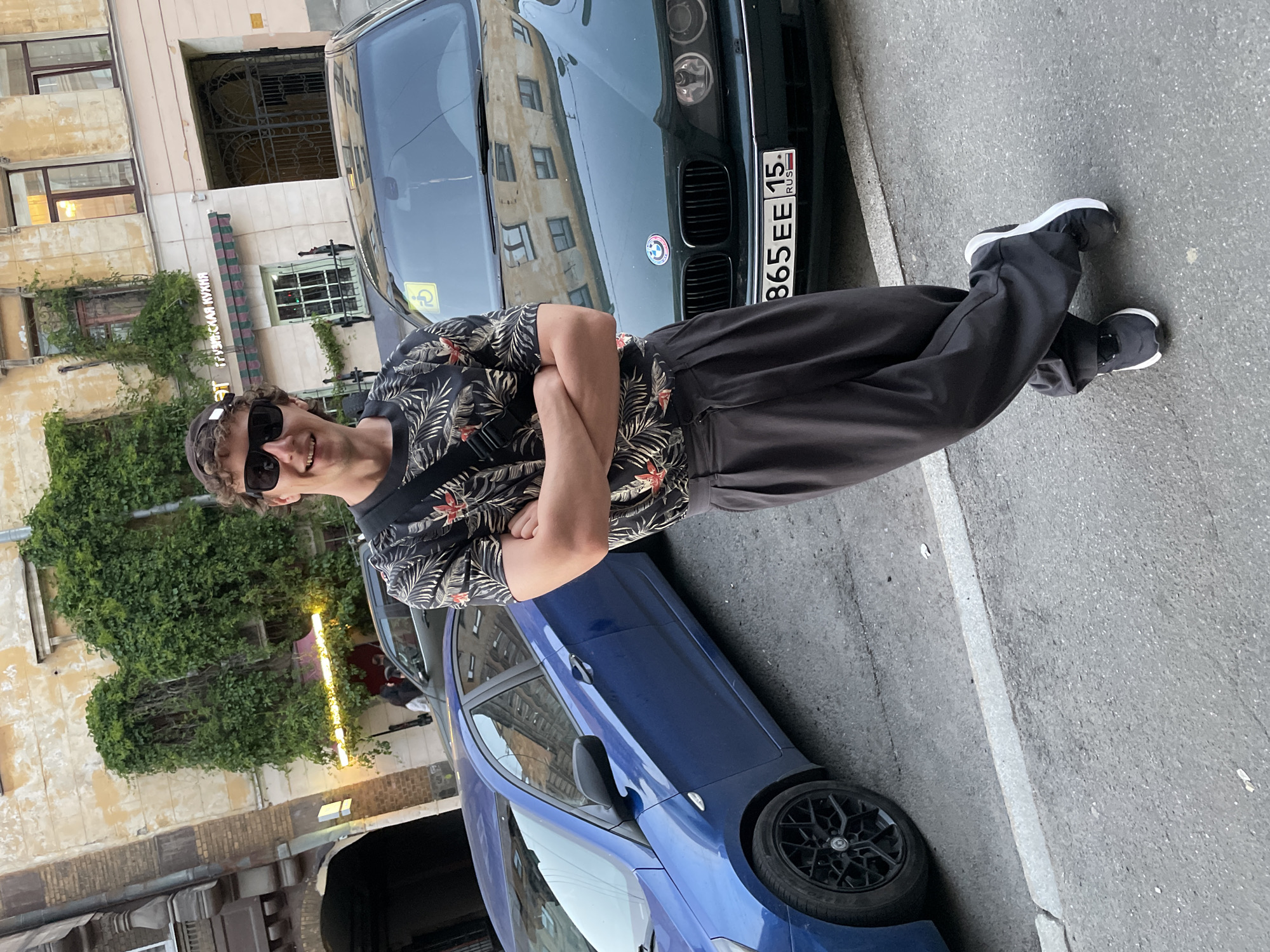 A young adult male stands centrally on an asphalt street, posing with a smile, arms crossed, and one foot crossed over the other, leaning slightly. He has curly light brown hair, wears sunglasses, a backward cap, a dark short-sleeved shirt with a red and green tropical floral pattern, wide dark grey trousers, and black and white sneakers. A black strap from a cross-body bag is visible across his chest.
Two cars are parked behind him. To his left is a blue Hyundai sedan with black multi-spoke wheels. To his right is a dark green BMW E39 5-series sedan, displaying a Russian license plate "B 865 EE 15 RUS" and a yellow handicapped sticker on its windshield.
The background features aged, multi-story buildings with faded yellow-orange facades, extensively covered in green climbing vines around windows and walls. An arched passageway is visible to the far left. A storefront sign in Cyrillic, reading "ГРУЗИНСКАЯ КУХНЯ" (Georgian Cuisine), is visible above a green and white striped awning on one building section. Below a section of heavy vine growth, a yellow neon light bar illuminates another storefront area. The architecture is consistent with older urban areas in Saint Petersburg, Russia.
