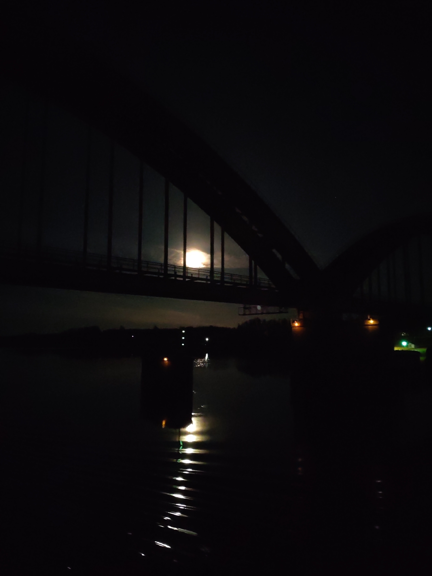 A dark, low-angle image depicts a bridge at night. The bridge's silhouette, a dark shape with vertical supports, arches over a body of water. A bright, round light source, possibly the moon, is visible through the bridge structure, partially obscured by clouds. The water reflects light in a series of bright, elongated patterns. Along the opposite bank of the water, a line of trees and bushes is visible in silhouette, punctuated by faint light sources, possibly streetlights or building lights. No people are visible. The location is unspecified in the image.