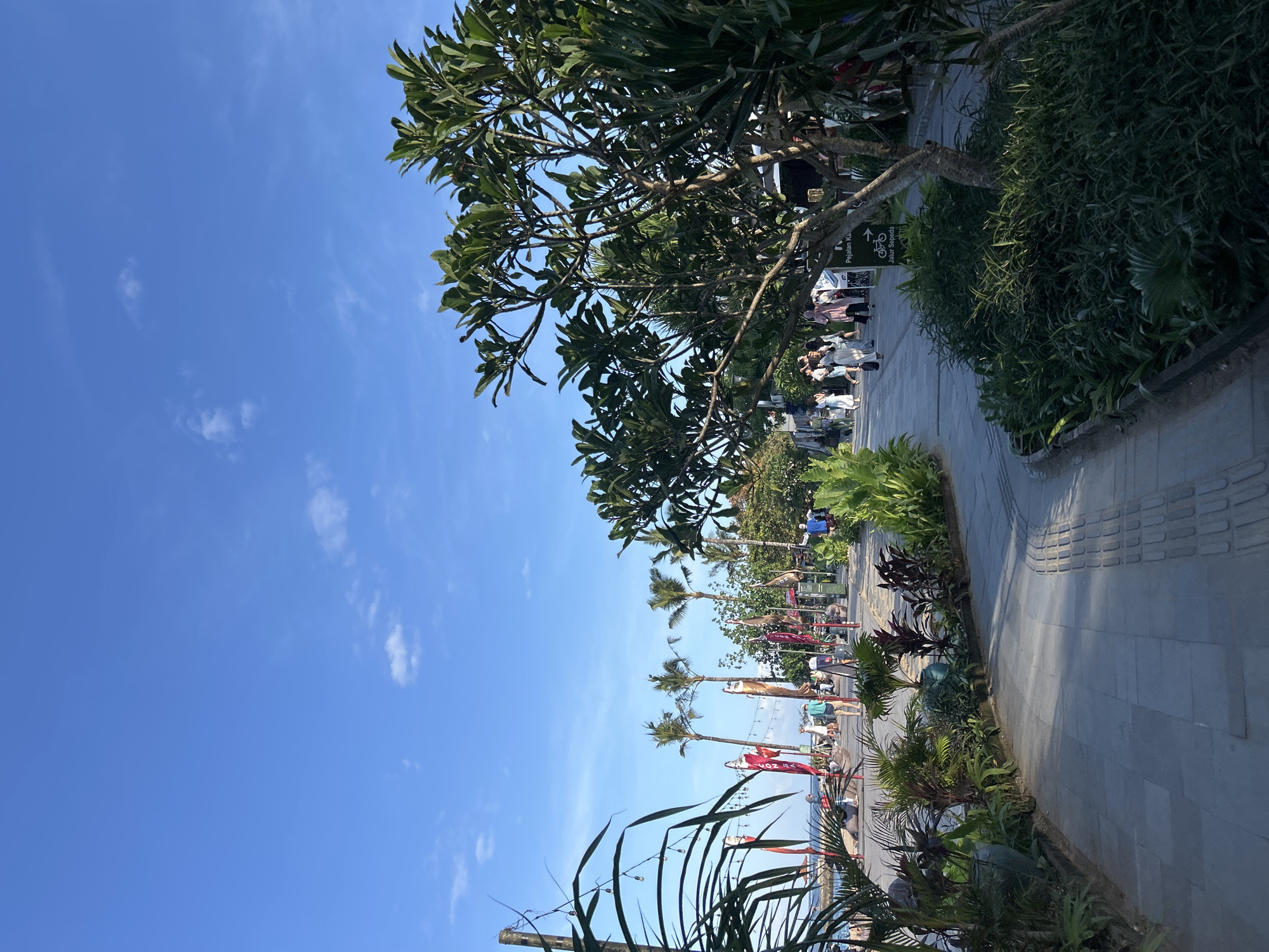 The image depicts a lively pedestrian walkway along a beach in Denpasar, Indonesia. The foreground features lush green foliage, including a large tree with dark branches and glossy leaves, bordering a paved pathway. The path itself is made of grey stone tiles and features tactile paving for visually impaired pedestrians. Beyond the walkway, a line of palm trees stretches across the background, with several people visible walking and engaging in various activities. Red and white flags are planted in the sand, adding a touch of color to the scene. In the distance, the ocean is visible, with a clear blue sky overhead dotted with a few wispy clouds. A signpost with Indonesian text is visible on the right side of the path.