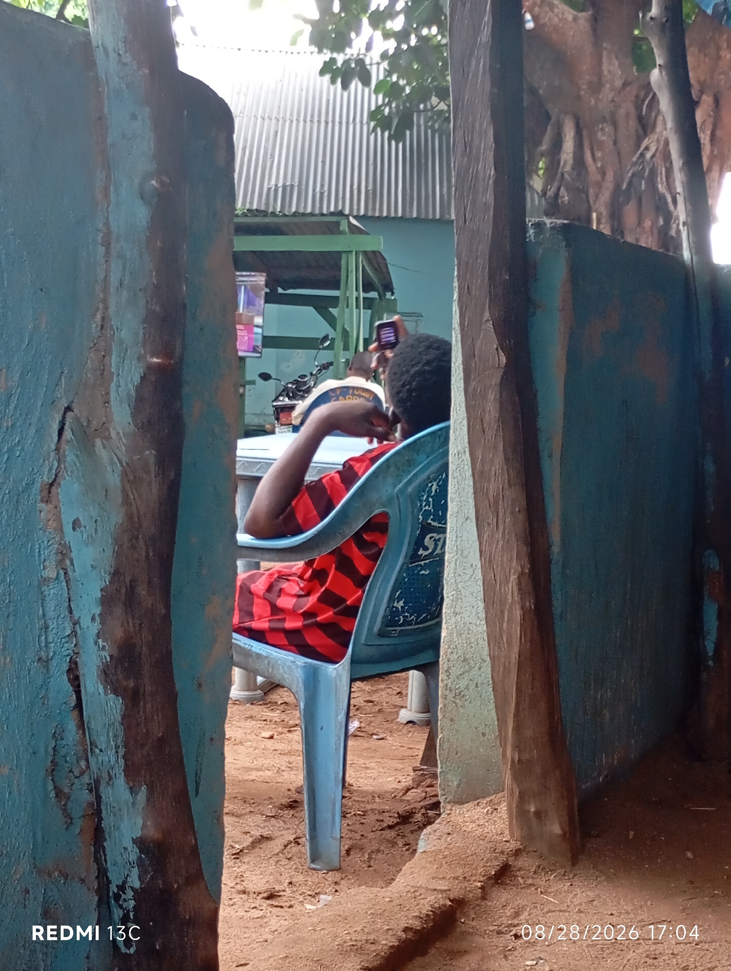 In Shendam, Nigeria, a person sits on a blue plastic chair, facing away from the camera. They are wearing a red and black striped shirt. The person is leaning back in the chair, with their left arm resting on the chair's armrest. They appear to be holding a phone in their right hand, near their face. The background shows a corrugated metal roof and the roots of a large tree. The ground is sandy and there are wooden posts in the foreground that are painted blue and brown. The image is dated 08/28/2026 at 17:04.