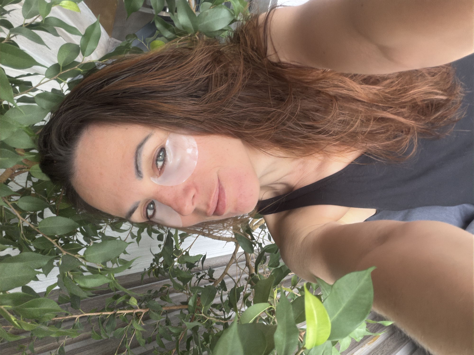 A person is positioned in front of a leafy plant. The person has fair skin, brown hair and is wearing eye masks. They are wearing a dark-colored top. The background consists of a light-colored wall or structure. The image is a selfie-style, close-up shot. There is no information in the image that supports a location in the city of Birobidzhan, Russia.