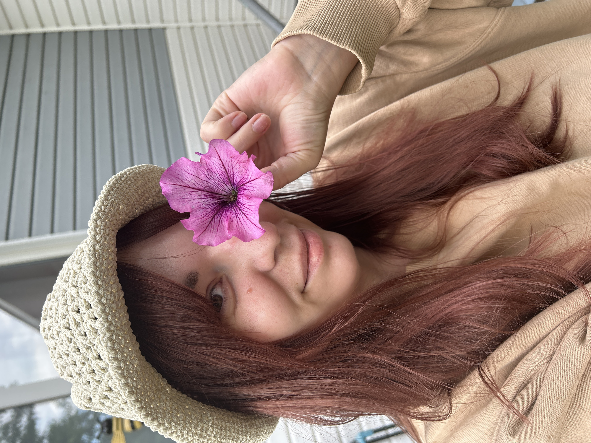 The image features a young woman, likely in Фоки, Russia, captured in a close-up, upward-angled shot. Her head is tilted, and she looks directly at the camera with a soft, slightly smiling expression. Her reddish-brown hair frames her face, flowing down her shoulders. She wears a light beige or tan hooded sweatshirt and a cream-colored, crocheted sun hat with a textured, open-weave pattern.
Her right hand, with natural nails, holds a single vibrant pink petunia flower to her face, partially covering her right eye. The flower's prominent dark purple veins are clearly visible.
The background consists of vertical, light grey paneled siding, suggesting an outdoor setting such as a building exterior, balcony, or porch. A small portion of what appears to be an overcast sky is visible in the upper left corner, indicating it is daytime. The overall lighting is soft and diffused. There is no visible text in the image.