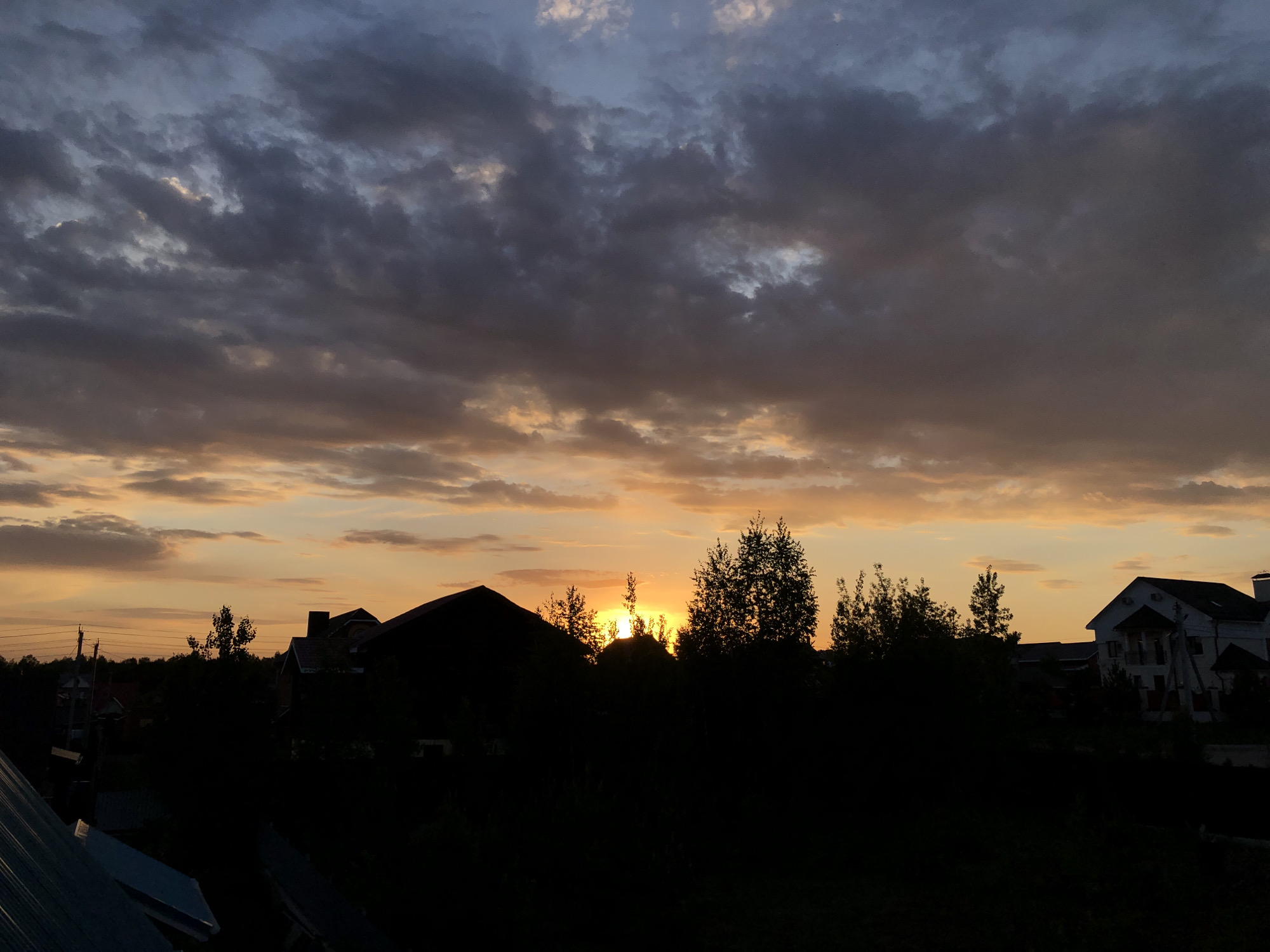 The image shows a sunset view in Noviy, Russia. The sky is filled with a mix of gray and orange clouds, with the sun partially obscured on the horizon. In the foreground, there's a silhouette of trees and buildings, including what appears to be residential houses. There are utility poles and wires visible in the distance to the left. The roofs of buildings are partially visible at the bottom of the frame.