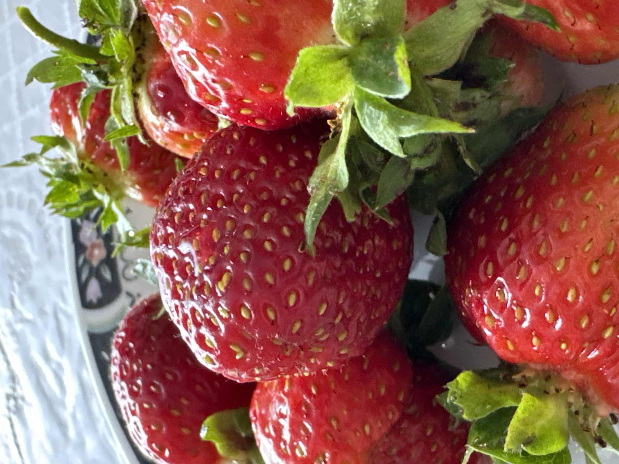 A close-up, overhead shot captures a bowl filled with ripe, red strawberries. The strawberries are plump and juicy, with visible seeds dotting their surfaces. Green leaves and stems are attached to some of the berries. The bowl itself has a decorative rim with a floral pattern. The background appears to be a textured white surface. The image does not depict any specific location in the city of Kumyony, Russia, nor does it contain any people or actions.