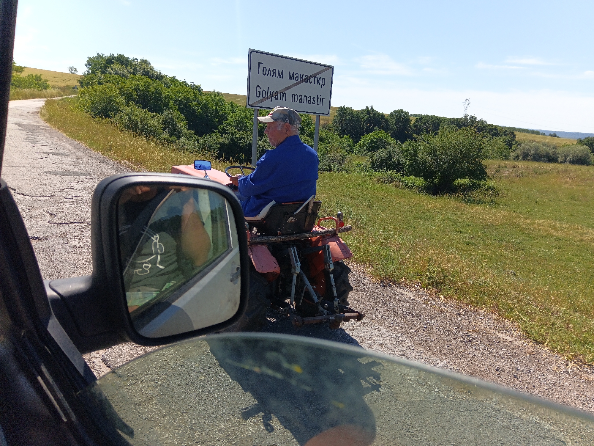 A man in a blue jacket and a camouflage baseball cap drives a red tractor on a paved road with gravel shoulders in a rural area. A sign in both Cyrillic and Latin script reading "Голям манастир Goly am manastir" is visible behind the tractor. The landscape is hilly and green, with trees and grass. The image is taken from inside a vehicle, with the side mirror and windshield reflecting the scene. The context is the village of Malak Manastir, Bulgaria.