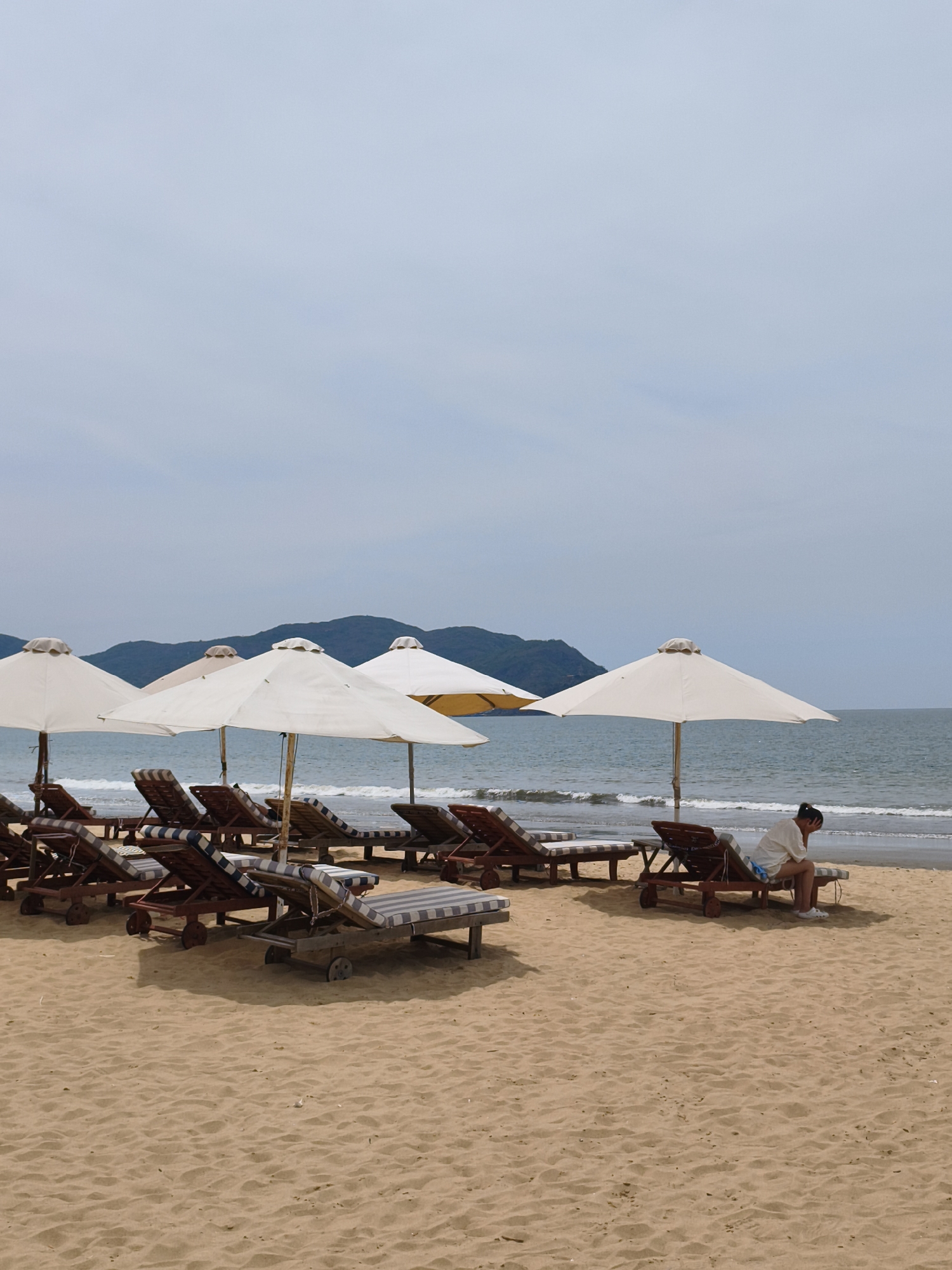 This is a photograph of a beach in Quy Nhon, Vietnam. The image depicts a row of empty lounge chairs and umbrellas set up on a sandy beach. The ocean is visible in the background with gentle waves. In the distance, there are green, tree-covered hills under a cloudy sky. A single person, dressed in a white shirt and shorts, sits on one of the lounge chairs, facing away from the camera and looking towards the ocean. The person's head is lowered, and they appear to be resting or contemplating. The overall atmosphere of the image is calm and serene.