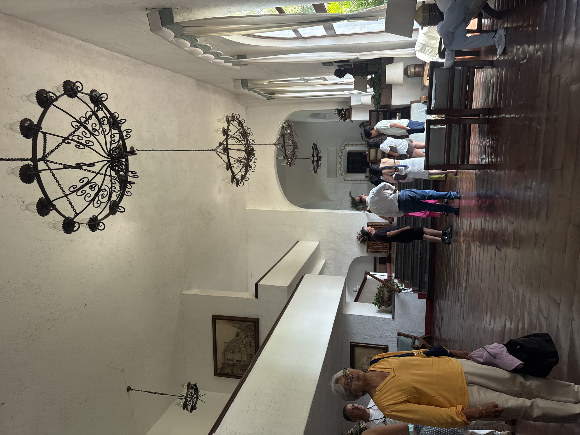 The image depicts the interior of a building, likely in Cocoyoc, Mexico, based on the architecture. A group of people are present, some standing in a line on a dark wooden floor, while others are positioned closer to the foreground. The architecture features white walls, arched doorways, and decorative molding. Ornate, wrought-iron chandeliers hang from the ceiling. A staircase is visible, leading to a higher level, and framed artwork adorns the walls along the stairway. Seating and decorative plants are also present within the space.
