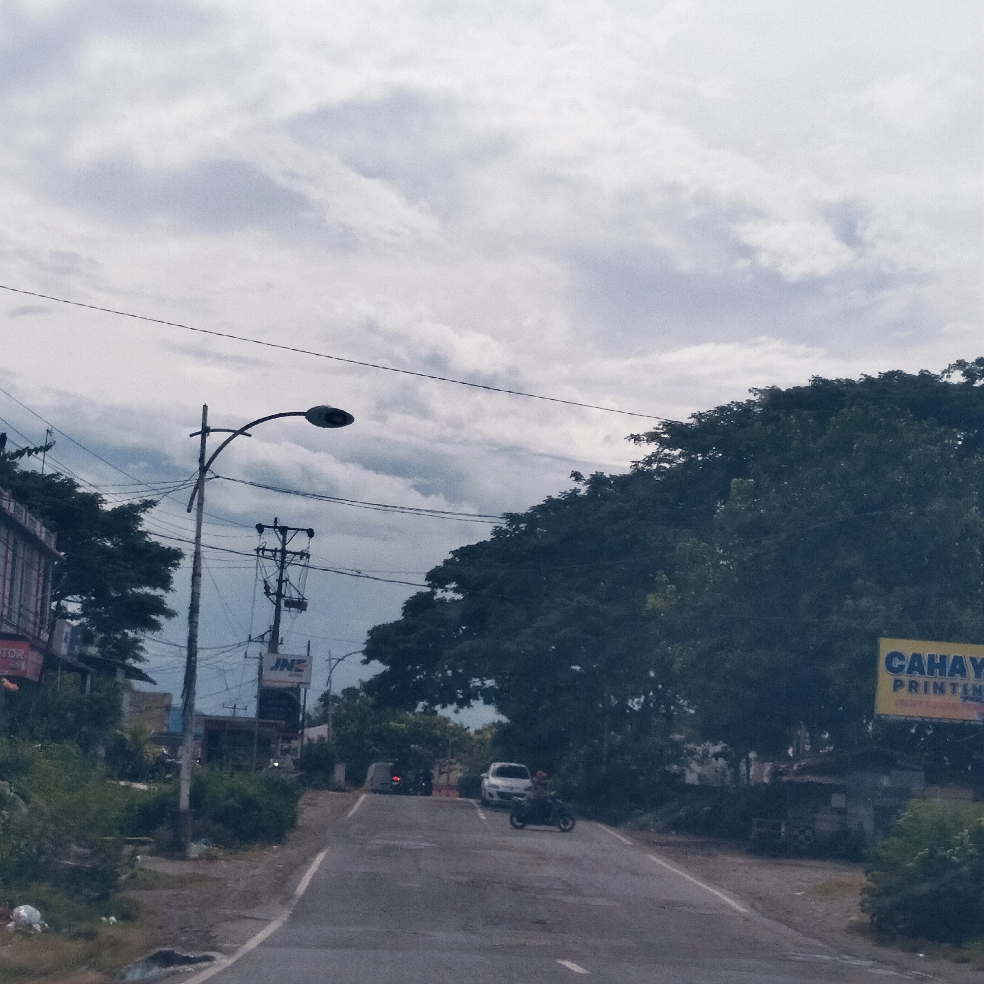 The image displays an overcast road scene in Cot Teungoh, Indonesia, depicting a quiet, semi-urban or rural environment. A narrow asphalt road, marked with a white line on its right edge, leads uphill into the distance. In the mid-ground, a person rides a motorcycle away from the viewer, while a white car is visible further ahead, and another vehicle is faintly discernable in the far distance.
The landscape is characterized by dense, dark green trees and bushes flanking both sides of the road. On the left, utility poles and a street lamp stand tall, intricately laced with numerous power lines against the cloudy sky. Partial views of buildings are on the left, with some visible text possibly reading "TOP" and "MOTOR." A white sign displaying the "JNE" logo (an Indonesian logistics company) is also visible on a pole on the left.
To the right, a prominent yellow billboard catches the eye, clearly displaying the text "CAHAYA PRINTING." Beneath the billboard and behind the trees, a small, rustic structure is partially visible. The sky is predominantly covered in soft grey and white clouds, indicating an overcast day without direct sunlight, contributing to a muted and tranquil atmosphere. No specific emotional expressions are discernable from the subjects.