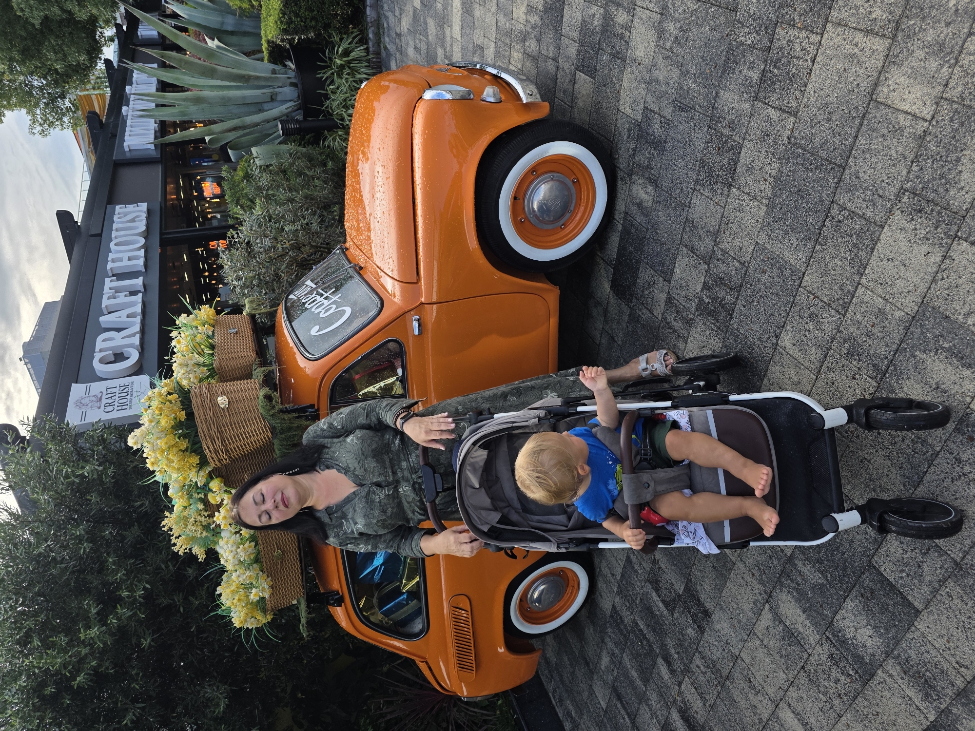 A woman with dark hair and closed eyes stands next to a vintage orange car with baskets of yellow flowers on its roof. A toddler sits in a stroller beside her, looking to the right. The car is parked on a brick-patterned walkway in front of a building with a sign that reads "CRAFT HOUSE." The background includes lush greenery and large agave plants. The scene appears to be outdoors, possibly in a commercial or public area. The car has white-walled tires and the word "Copper" is visible on the windshield. The sky is overcast, and there are some water droplets on the car's surface, suggesting it may have rained recently.