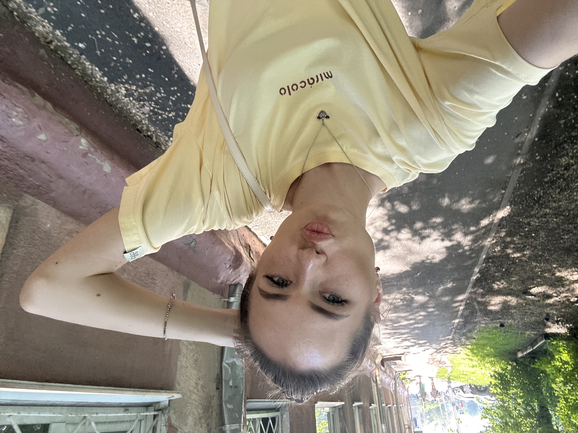 A young woman with her hair pulled up is taking a selfie outdoors on a sunny day. She is wearing a pale yellow t-shirt with a necklace and a bracelet. Her left arm is raised behind her head, and she is puckering her lips. The background shows a tree-lined sidewalk next to a building. The sidewalk is dappled with sunlight and shadow, and there are some fallen leaves on the ground. A few people and cars are visible in the distance.