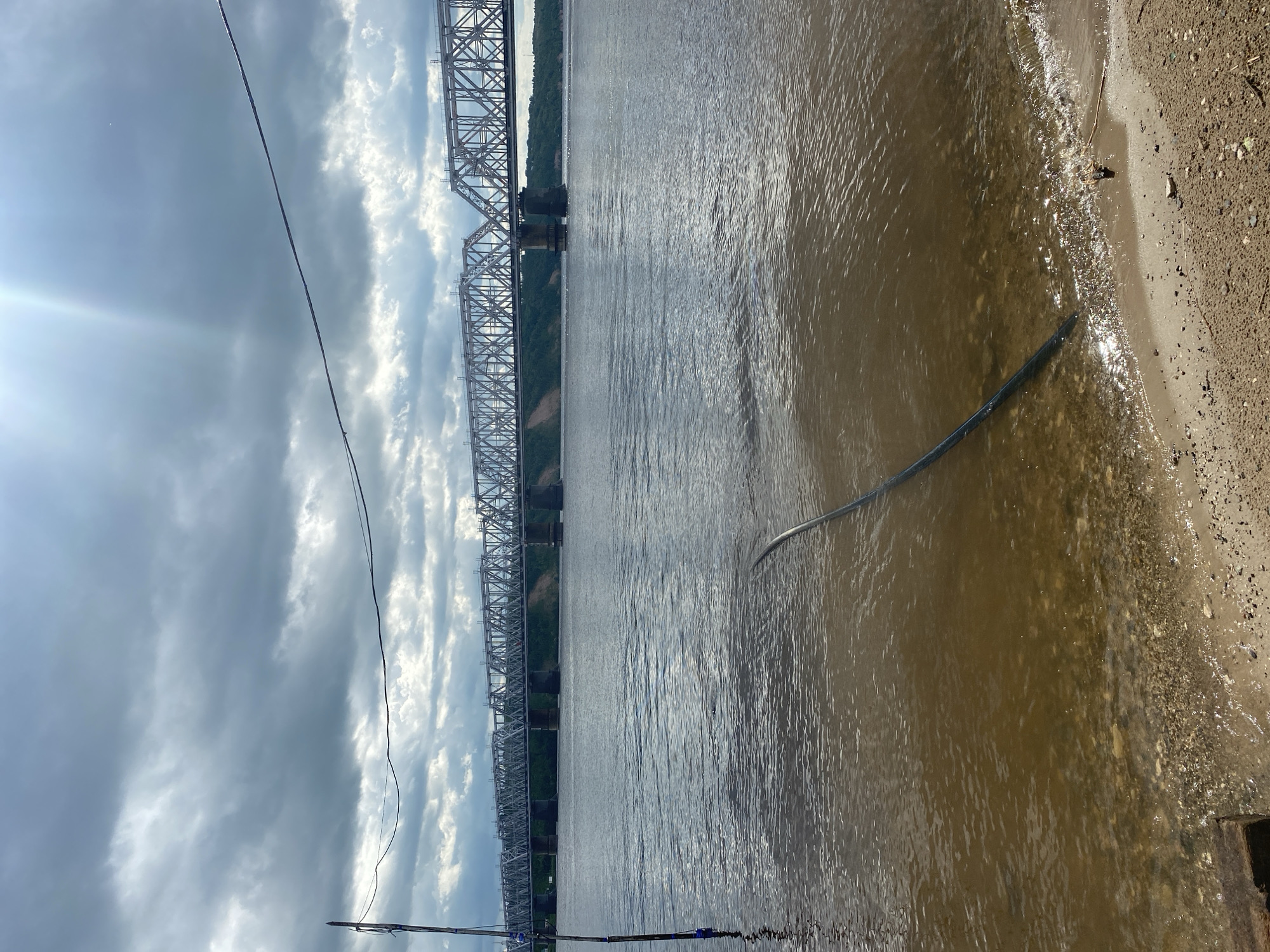 The image depicts a body of water, likely a river, under a cloudy sky. A large, silver metal truss bridge spans across the water, stretching from the left side of the frame towards the right background. The bridge appears to be carrying a railway line. On the right side of the image, there is a sandy bank with some debris and a dark, curved object, possibly a pipe or hose, partially submerged in the water near the shore. In the sky, there are dramatic, white clouds against a blue-grey background, suggesting overcast or rapidly changing weather. Sunlight pierces through the clouds on the far left, creating a lens flare effect. A few power lines are visible in the foreground, stretching across the frame from bottom left to upper right. The water is choppy with visible ripples and reflections, indicating some movement or wind. The scene appears to be set during the daytime, given the lighting conditions and sky. The location is identified as Zelenodol'sk, Russia, which implies the bridge is a significant piece of infrastructure in that area, likely a crossing over a substantial river. There is no visible text in the image. The overall mood is somewhat dramatic due to the sky and the imposing structure of the bridge.