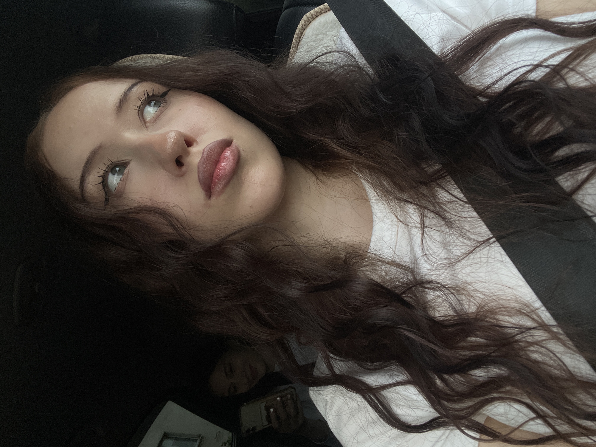 A young woman with long, wavy brown hair is captured in a close-up shot, looking to her right. She is wearing a white t-shirt and a black seatbelt is visible across her chest. Her makeup includes winged eyeliner and lipstick. In the background, another person is visible in a car, holding a smartphone. The car appears to be in motion, suggested by the slightly blurred background. There are no explicit indicators of the city of Votkinsk, Russia.
