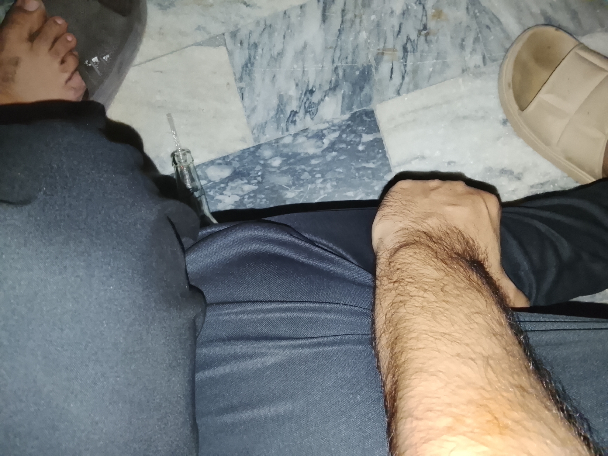 The image shows a first-person perspective of a person seated indoors in Shalimar Colony, Pakistan. The person is wearing gray sweatpants and has a tan sandal on their right foot and a gray slide on their left foot. A glass bottle with a straw is on the floor near the left leg. The floor is covered with gray and white marble tiles. The person's right forearm, which is hairy, is visible in the lower right portion of the image.