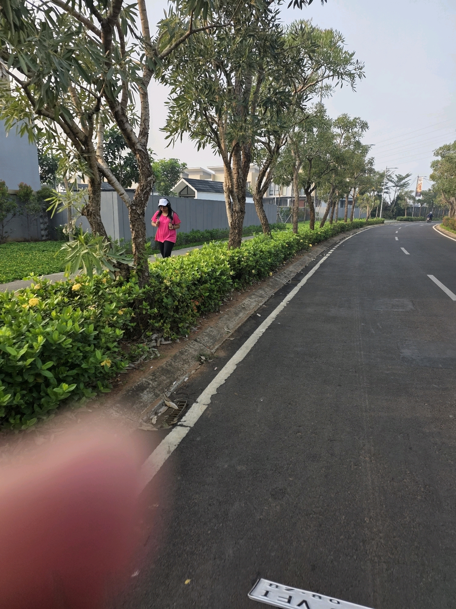 In the city of Bojongsoang, Indonesia, a woman wearing a pink shirt, black leggings, and a white cap jogs on a sidewalk adjacent to a road. She is carrying a backpack. The sidewalk is lined with lush green bushes and trees. The road is paved and has white lane markings. In the background, houses and power lines are visible. The scene appears to be captured in the morning or late afternoon, given the soft lighting. A portion of a red object, likely a finger, is blurred in the foreground, obscuring a part of the image. A license plate is also visible on the road.