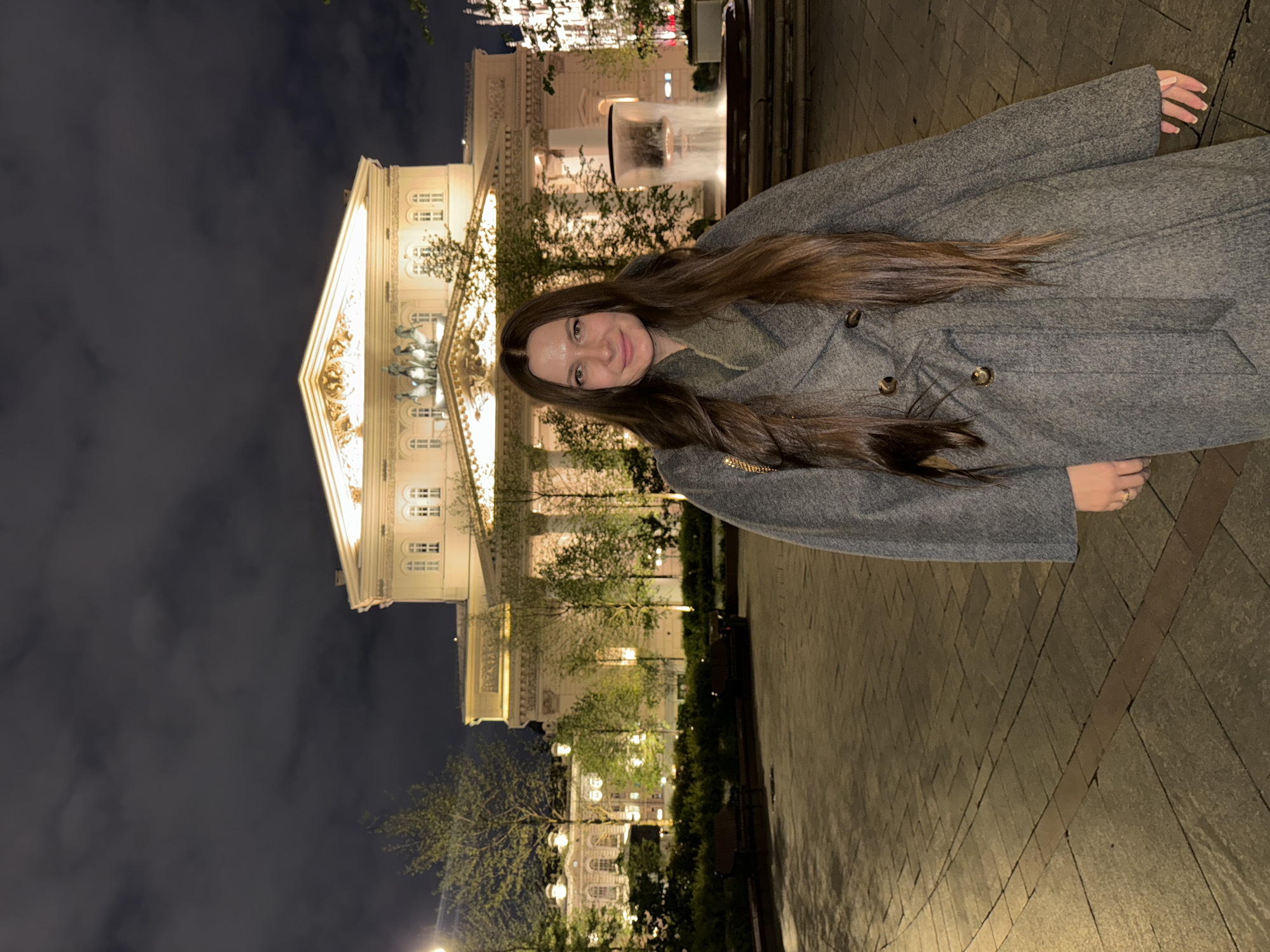 A woman with long brown hair, wearing a gray overcoat, stands on a cobblestone walkway. Behind her is the Bolshoi Theatre in Moscow, Russia, illuminated at night. A fountain is visible near the theatre, and trees line the walkway with lit street lamps. The sky is dark.