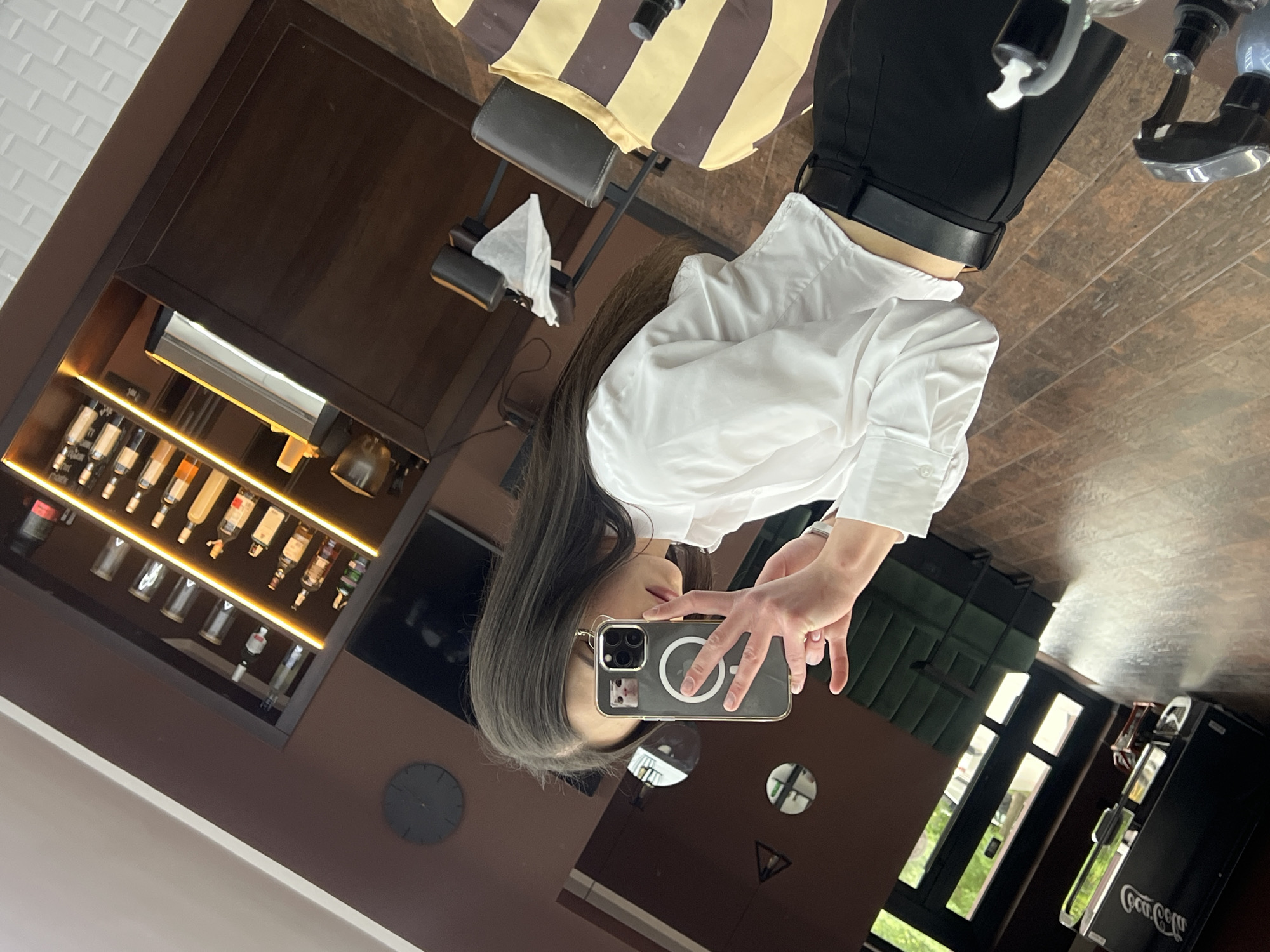 A person is taking a mirror selfie in what appears to be a barbershop or salon, with a barber's chair visible behind them. The person is wearing a white shirt and black pants. The shop has a dark wood interior. There is a backlit display of bottles, and a Coca-Cola vending machine in the background. A clock on the wall is visible in the mirror. The reflection implies the setting is in Ternopil, Ukraine.