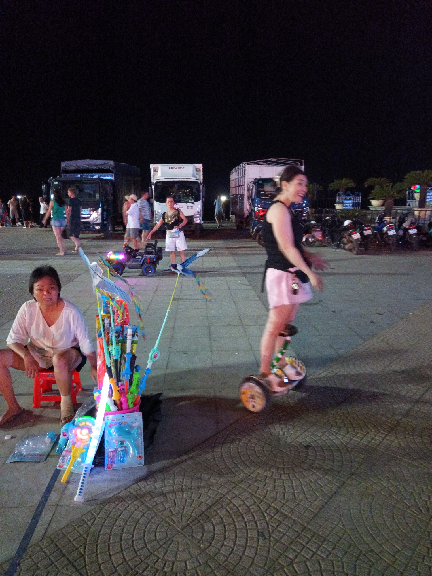 At nighttime on a paved area in Nha Trang Ward, Vietnam, a woman with short hair is seated on a small red stool. She is wearing a light-colored shirt and shorts. In front of her is a collection of colorful toys, including bubble guns, light-up swords, and kites. To her right, a woman is riding a hoverboard, wearing a black tank top and a light pink skirt. She is smiling and looking towards her left. In the background, several large trucks are parked, and a small electric toy car is visible. Motorcycles are also parked in the background. The ground is paved with interlocking circular and square stone patterns.