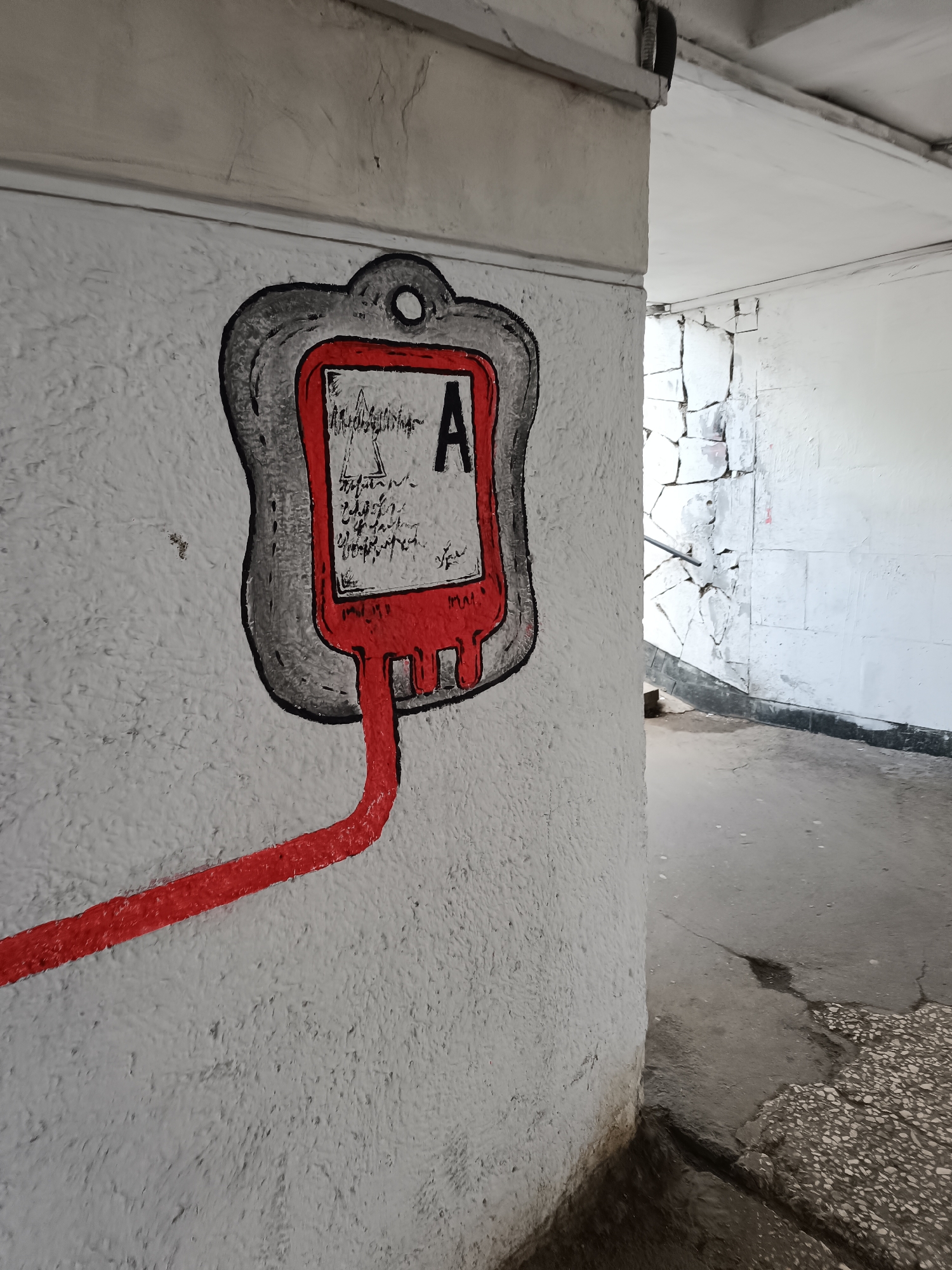 The image depicts a wall with a graffiti-style painting of a blood bag. The blood bag is labeled "A" and is attached to a red tube extending down the wall. The wall is textured and painted white. To the right of the wall is a passage or hallway leading into an unknown area. The floor is cracked and appears to be made of concrete. The location is Unknown, Unknown.