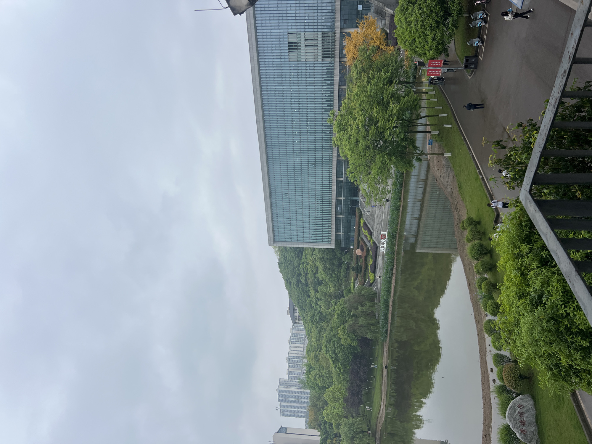 The image shows a wide-angle view of an outdoor scene in Jiangjin, China, captured on an overcast day. The focal point is a large, modern glass building on the left, reflecting the sky and surrounding greenery. In front of the building is a body of water reflecting the trees and buildings. A road and pathway run along the right of the building, with trees, bushes, and grass. A few people are walking on the road. On the far left and behind the building is a hill covered with trees. Further left, in the distance, are high-rise apartment buildings. A metal railing is visible on the right.