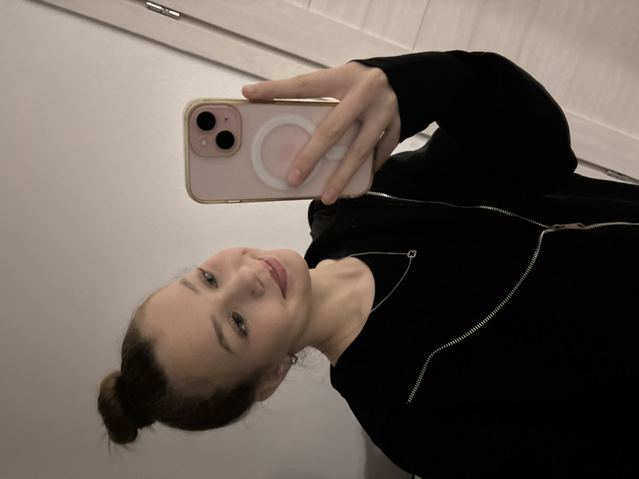 A young woman takes a selfie. She is holding a pink iPhone with a white ring on the back. She wears a black zippered jacket with an asymmetrical zipper. Her hair is pulled back in a bun. She is facing forward and slightly tilted, smiling softly. The background appears to be a white or light-colored wall.