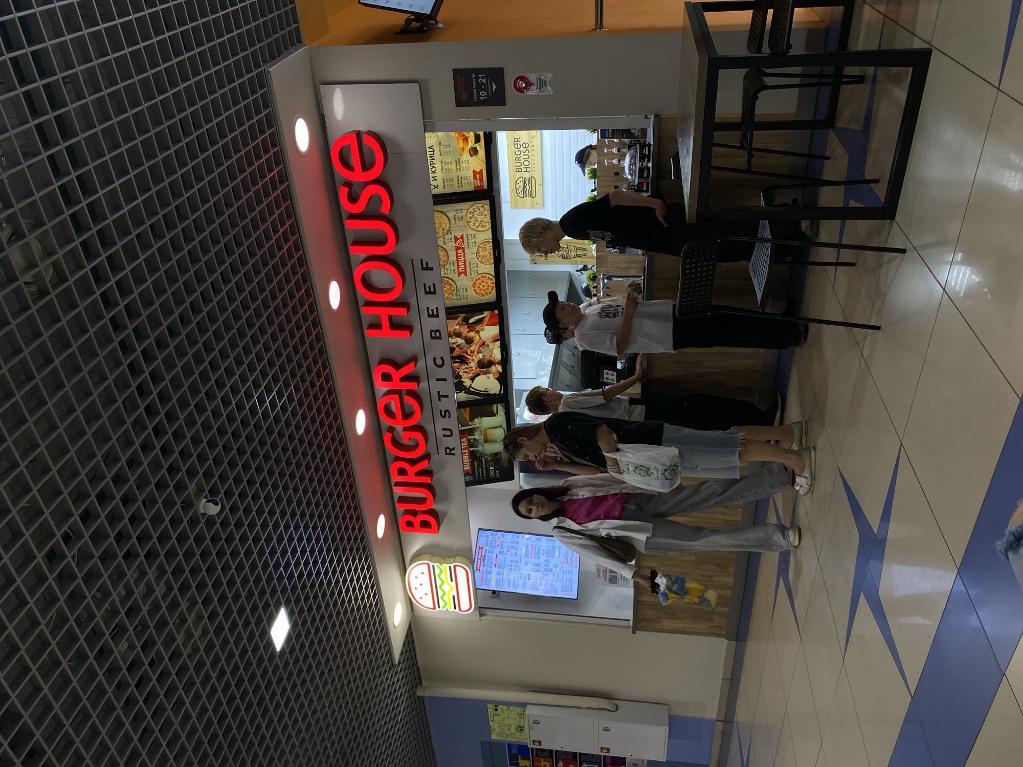 The image shows the interior of a Burger House restaurant, a fast-food establishment in Votkinsk, Russia. The main subject is a group of young people standing in line to order food. The restaurant's signage is prominent, with large red "BURGER HOUSE" lettering and various menu boards displaying food items like pizza and chicken.
The scene is brightly lit, suggesting it's daytime, and the interior appears to be a typical mall food court setting with tiled flooring featuring a blue star pattern. A table with chairs is visible in the foreground, suggesting a dining area. The people in line are casually dressed, and their expressions are neutral, indicating a routine food ordering activity. One person in line is looking at the menu on a digital screen.
Visible text includes the restaurant name "BURGER HOUSE," menu items like "ПИЦЦА" (Pizza), "И КУРИЦА" (and Chicken), "КРЫЛЬЯ СТРИПСЫ НАГГЕТС" (Wings, Strips, Nuggets), and "BUBBLE TEA." There is also text indicating the operating hours: "РЕЖИМ РАБОТЫ 10-21" (Opening Hours 10-21). A small sign with a burger logo and the founding year "SINCE 2013" is also present. The overall impression is a casual, everyday scene at a popular fast-food chain.