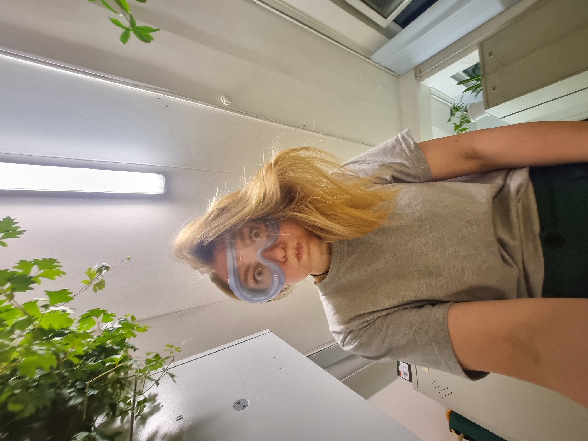 A young person with blonde hair is wearing safety goggles. They are looking down at the camera. A green leafy plant is visible to the left. A white, rectangular light fixture is visible on the ceiling. There is a white cabinet or appliance with a circular opening on its surface. The setting appears to be indoors, possibly a laboratory or a room with plants. The location in the city of Kemul, Russia, is unknown.