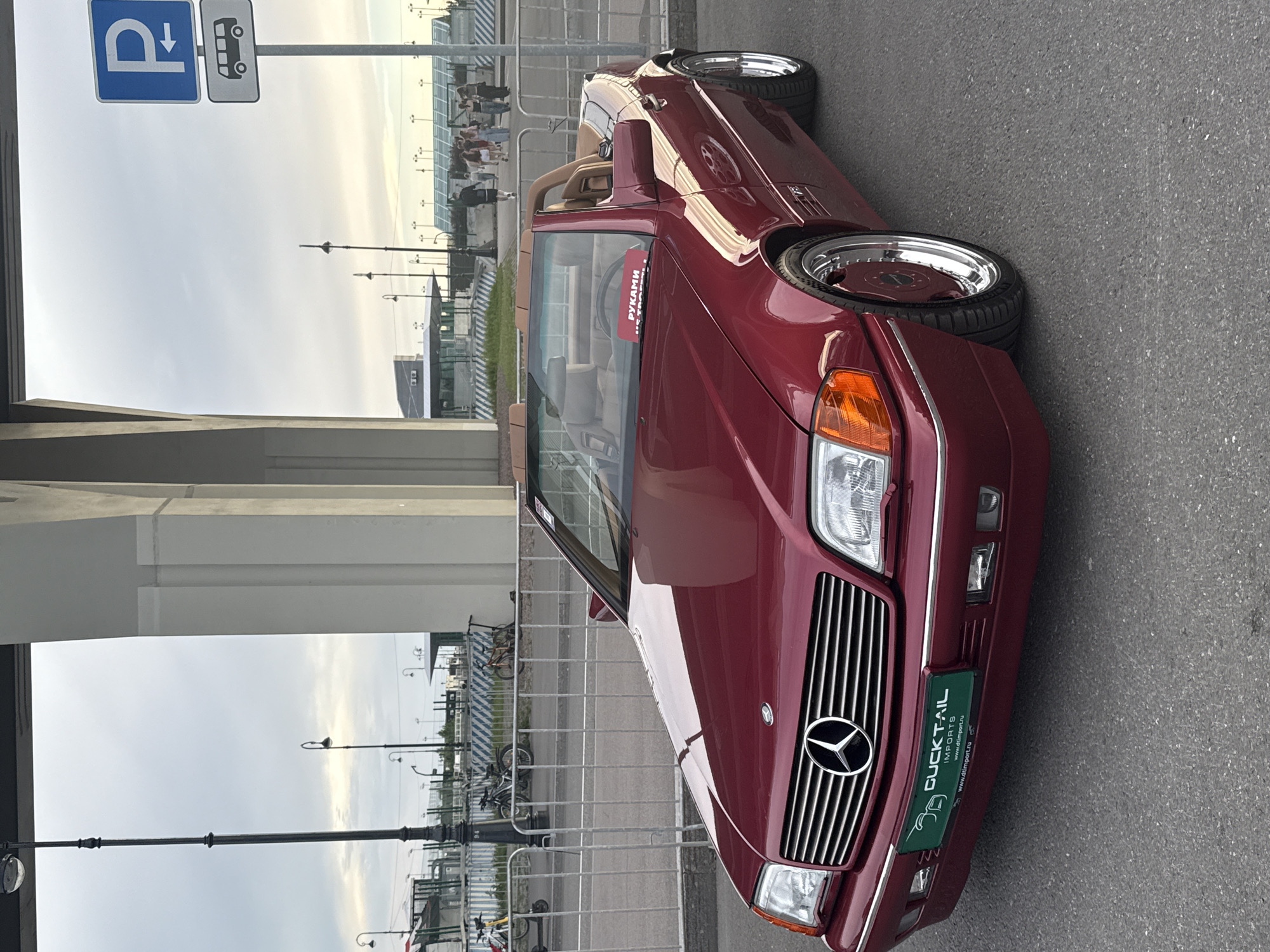 The photograph captures a maroon Mercedes-Benz SL convertible parked on asphalt, with a grey metal fence in the foreground. Behind the fence, several bicycles and scooters are visible, along with lampposts. In the background, a modern building with a glass roof and a group of people walking can be seen. To the right, a blue and white parking sign indicates a bus parking area. The sky is overcast. The car is adorned with a green license plate that reads "DUCKTAIL IMPORTS" and "www.d4import.ru". A small red sign on the dashboard reads "РУКАМИ НЕ ТРОГАТЬ". The scene appears to be an outdoor event or gathering. The architectural style of the overpass and the overall environment are consistent with the area near the Tauride Garden or potentially the area around the Saint Petersburg Stadium, both located in Saint Petersburg, Russia.