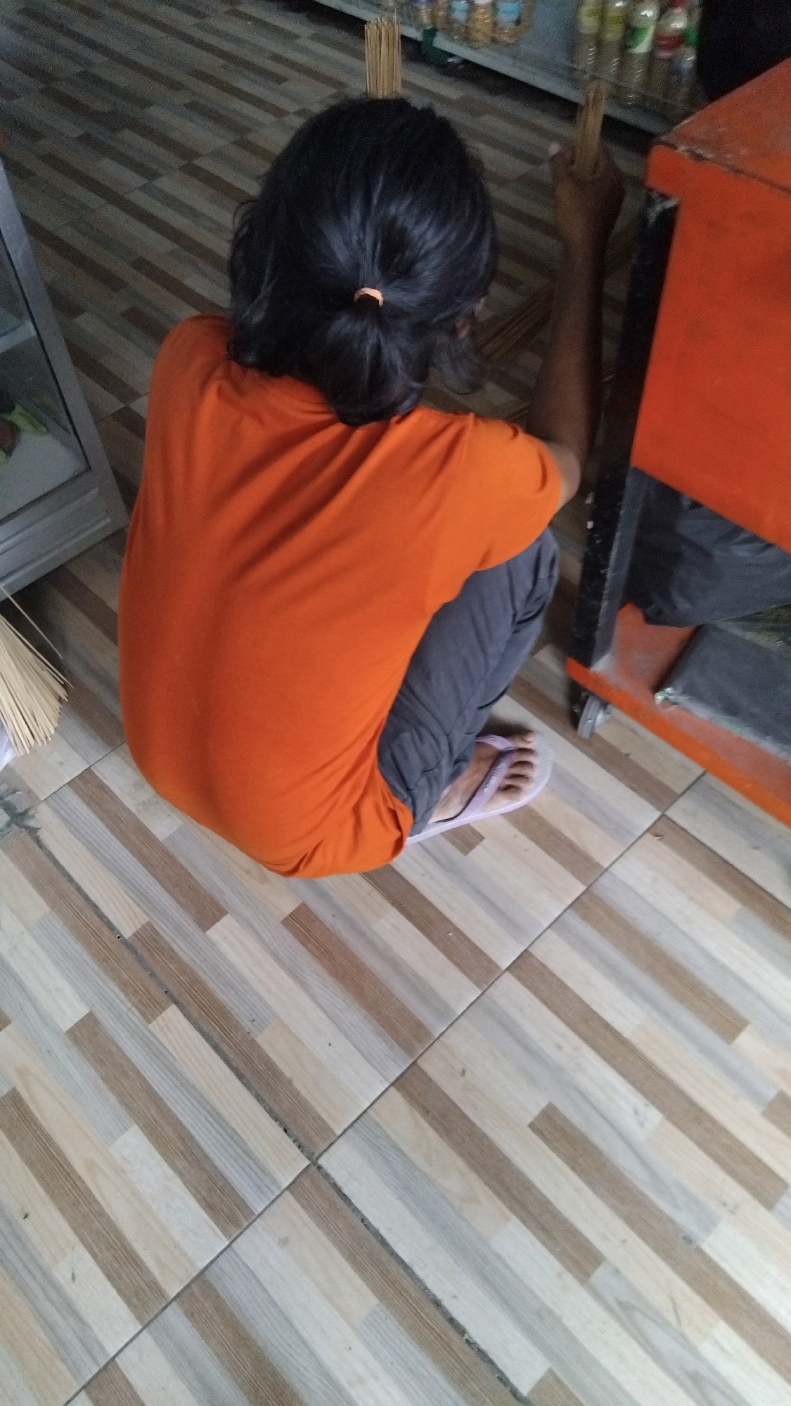 A person with dark hair pulled into a low ponytail is crouching on a tiled floor. They are wearing an orange t-shirt and dark pants, with light purple flip-flops on their feet. The person is reaching towards an orange shelf unit. Behind the person and to the left, a glass display case is partially visible. The floor tiles are light brown and beige, laid diagonally. The scene appears to be an indoor commercial space, potentially a shop, in Tegal, Indonesia, given the context.