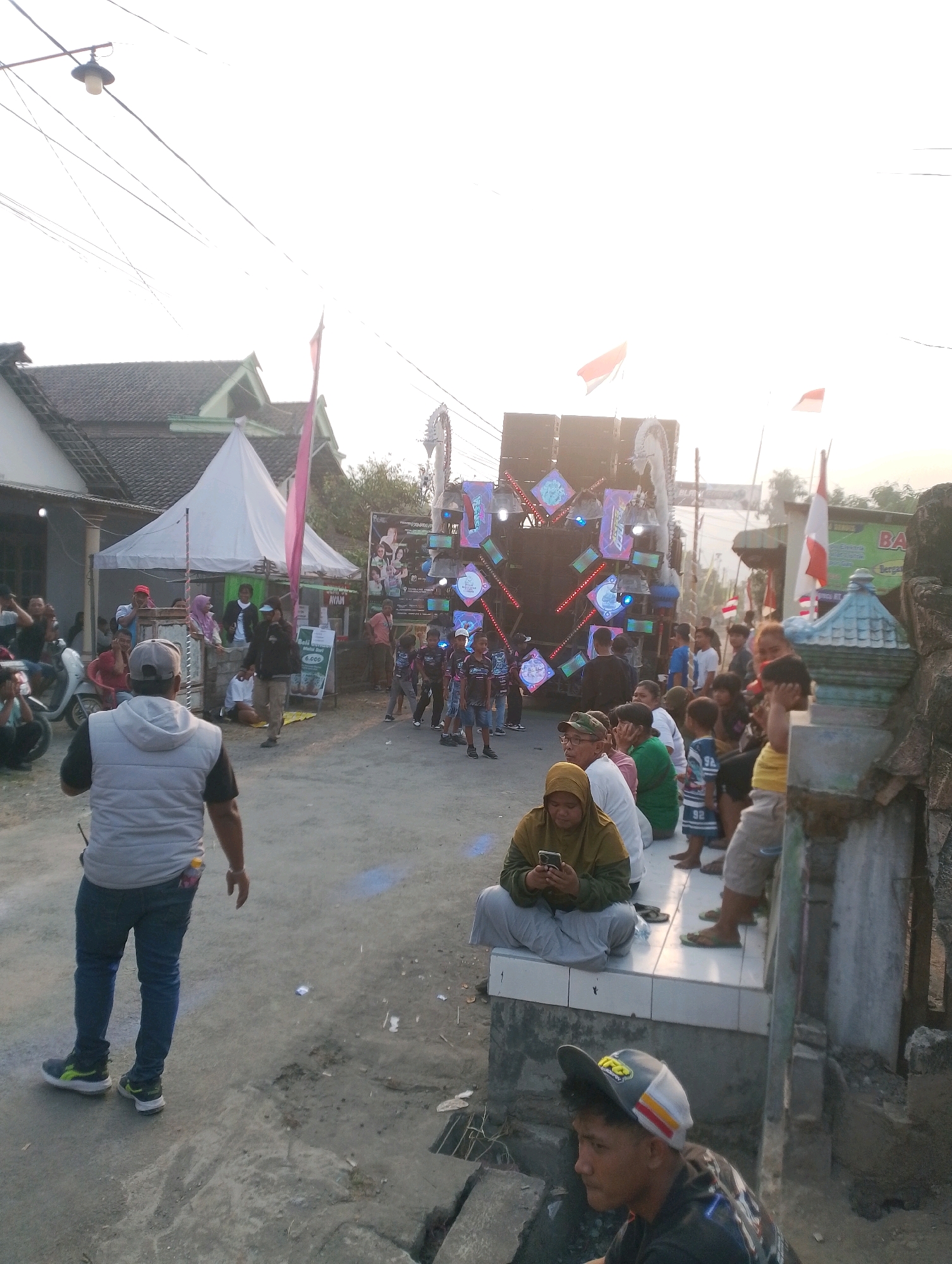 The image depicts an outdoor gathering, likely a community event, in a street in Rembang, Indonesia. The scene is dominated by a large sound system set up on a truck or platform, decorated with colorful lights and banners. Several people are gathered around, with some individuals seated on a raised concrete platform on the right side of the image. A woman in a yellow headscarf is using her smartphone while seated. Children are visible in the foreground and middle ground, some appearing to be watching the event. A man in a grey vest and jeans is walking away from the camera on the left side of the street. Flags, including Indonesian flags, are displayed, suggesting a national or local celebration. The environment appears to be a residential street with houses visible in the background. The lighting suggests it is either early morning or late afternoon.