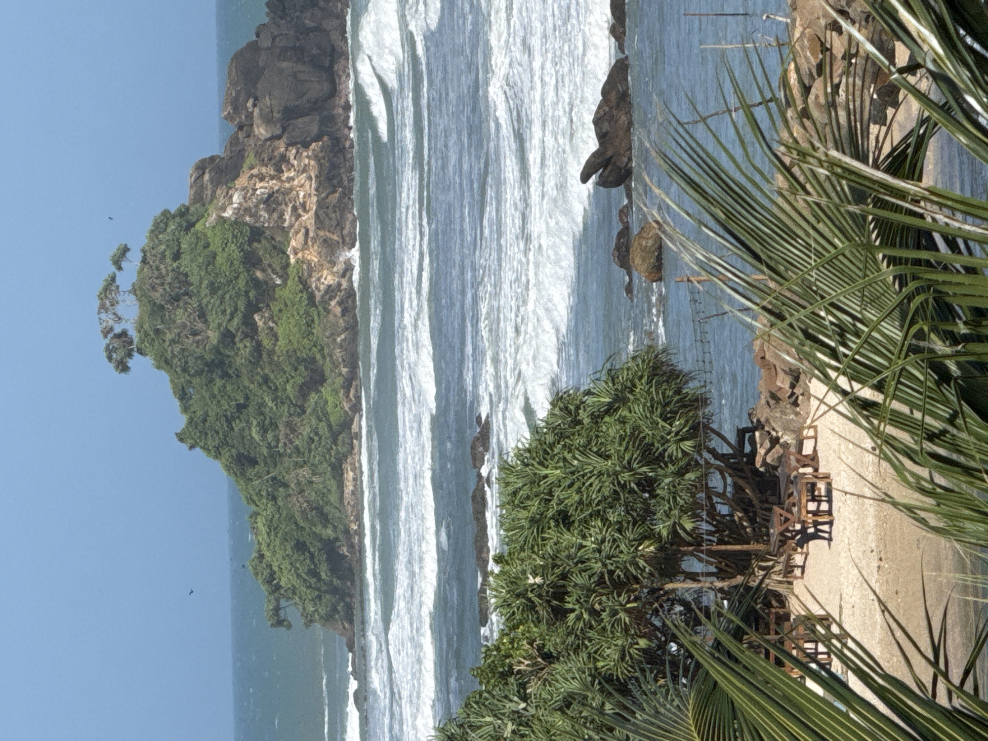 The image captures a scenic coastal view in Goviyapana, Sri Lanka, characterized by a small, verdant island rising from the ocean, surrounded by white-capped waves under a clear blue sky. In the foreground, a sandy beach area is dotted with several wooden tables and benches, shaded by lush green foliage. Palm fronds frame the lower part of the image, adding to the tropical ambiance. The ocean stretches to the horizon, with rocky outcrops visible in the water, and a larger, tree-covered island with rugged rock formations in the mid-ground. The overall scene depicts a tranquil beachside setting with natural beauty and relaxed outdoor seating.