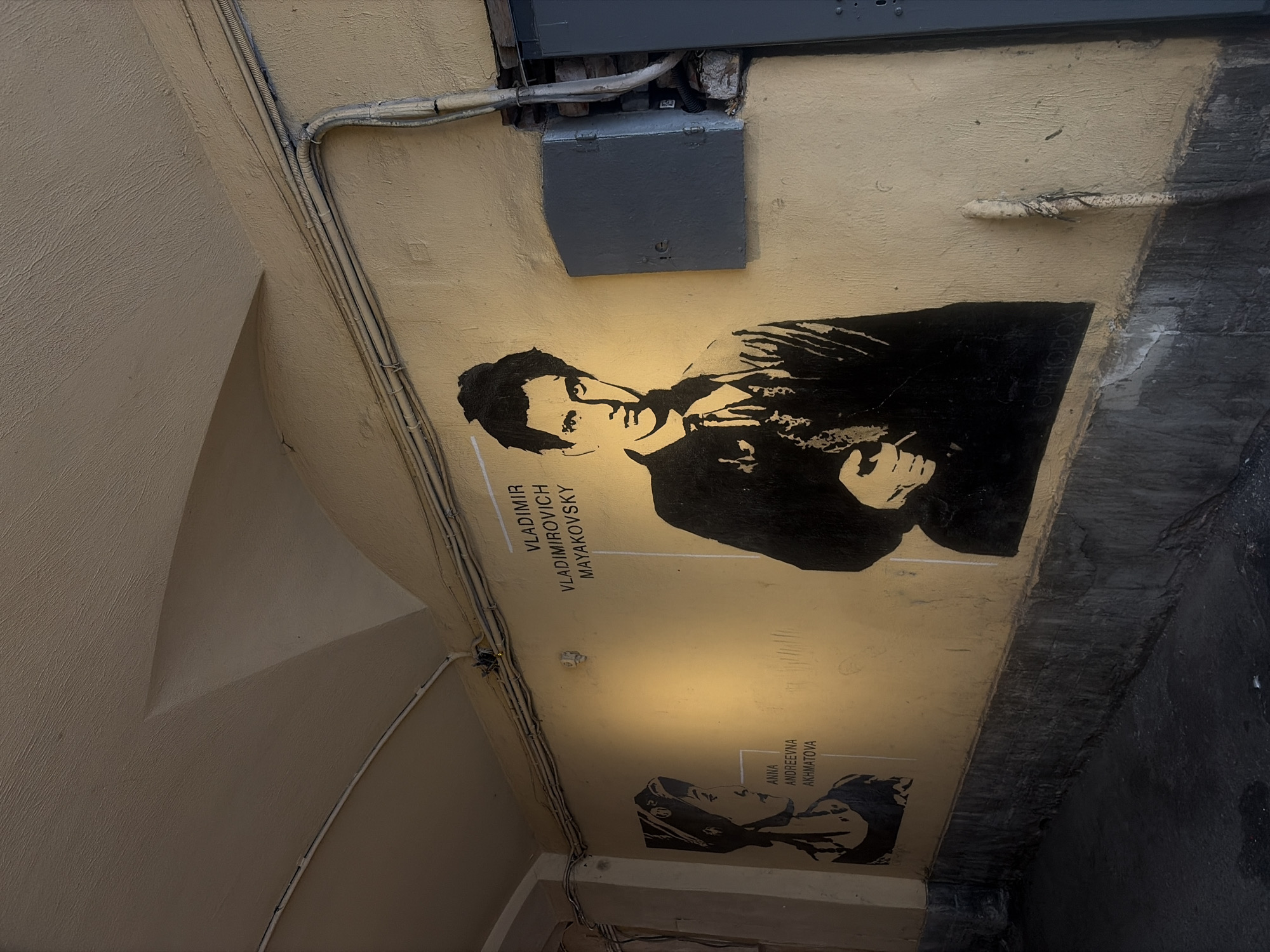 The image depicts a section of a building's exterior wall, likely in Saint Petersburg, Russia, judging by the style of the architecture and the names on the wall. The wall is painted a light yellow. Mounted on the wall are two large black and white stencils. The stencil on the right is of a man, identified by text above as "VLADIMIR VLADIMIROVICH MAYAKOVSKY." He is depicted from the chest up, looking directly at the viewer, wearing a suit jacket and tie. The stencil on the left is of a woman, identified by text beside her as "ANNA ANDREEVNA AKHMATOVA." She is shown in profile, wearing a headscarf.
Above the stencils, electrical conduits and wires run along the wall and across the arched ceiling. To the right of the Mayakovsky stencil, there is a grey electrical box attached to the wall, with some exposed brickwork and wiring visible beside it. The bottom of the wall is a dark, textured surface, possibly a stone base or a heavily weathered section, which transitions into a dark, asphalt-like ground. The overall impression is one of a public space, possibly an alleyway or courtyard, decorated with art referencing prominent Russian literary figures.