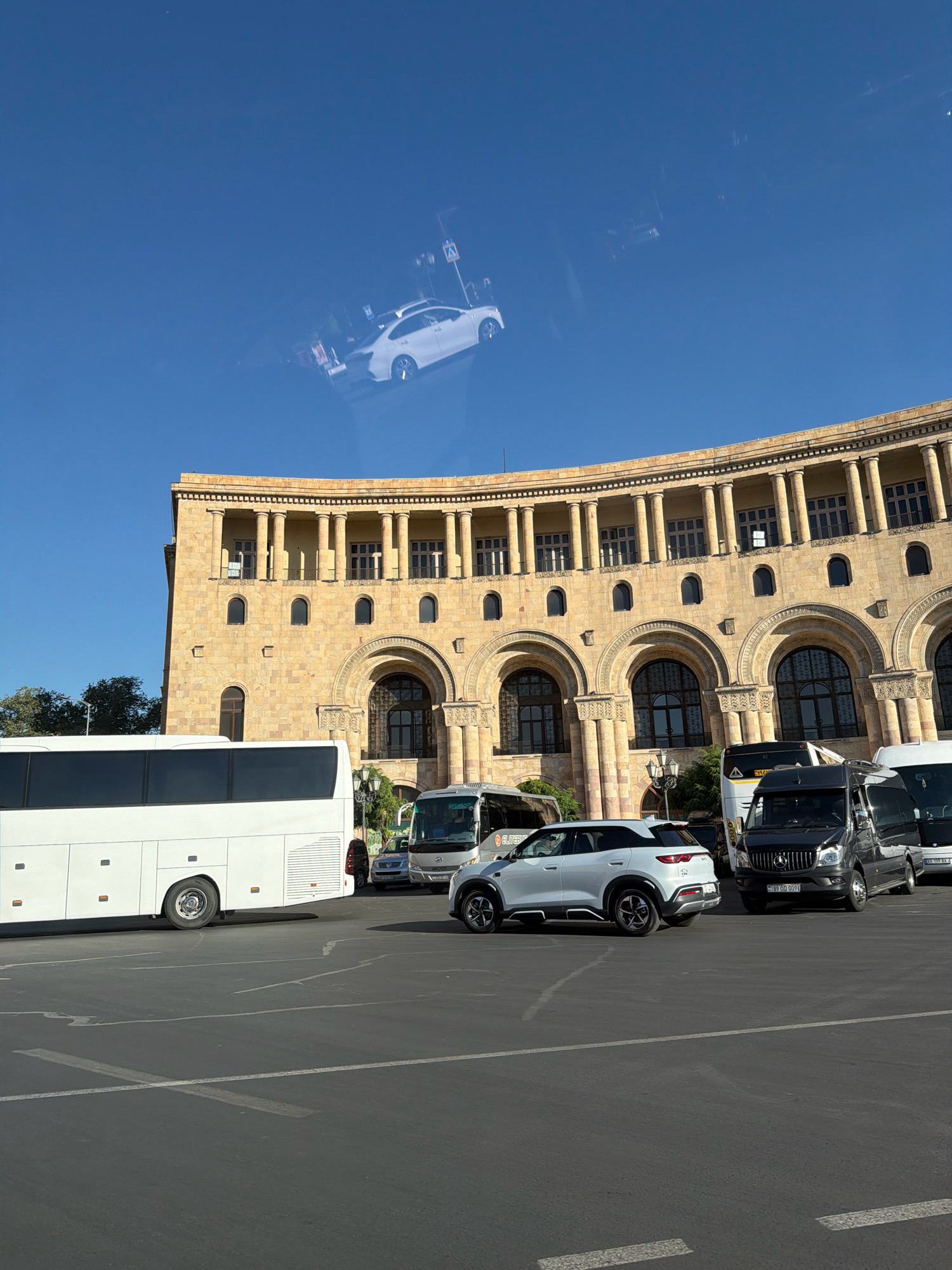 The image captures a sunny day in Yerevan, Armenia, focusing on the iconic Republic Square. A large, curved, sandstone building with classical architecture, characterized by columns and arched windows, dominates the background. In the foreground, several vehicles are parked on an asphalt lot, delineated by white parking lines. A long, white tour bus is parked on the left. To the right of the bus, a white SUV is positioned in front of a black Mercedes-Benz Sprinter van. Further to the right, another white van is visible. The sky is a clear, bright blue, reflecting the sky and some buildings, including a white car, on its surface. The overall scene depicts a typical day at a prominent public space in Yerevan.
