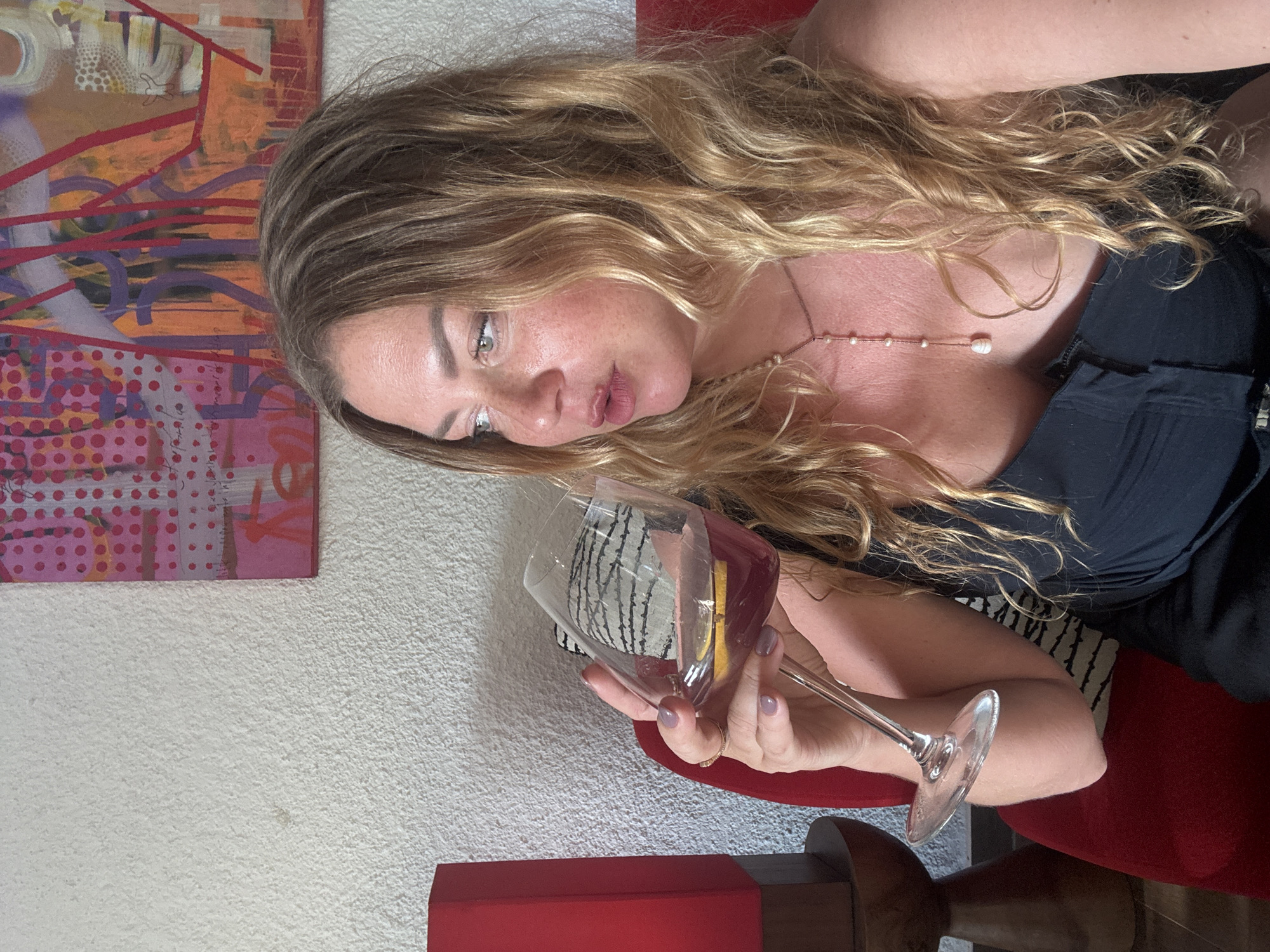 A woman with long, blonde, wavy hair is lying on a surface, holding a wine glass. The woman is looking at the wine glass with a thoughtful expression. The wine glass contains a pinkish-red liquid. She is wearing a dark-colored top and a necklace with small pearls. In the background, there is a painting with abstract designs and bold colors, and a white textured wall. The scene appears to be indoors, possibly in a home. The lighting suggests it could be daytime.