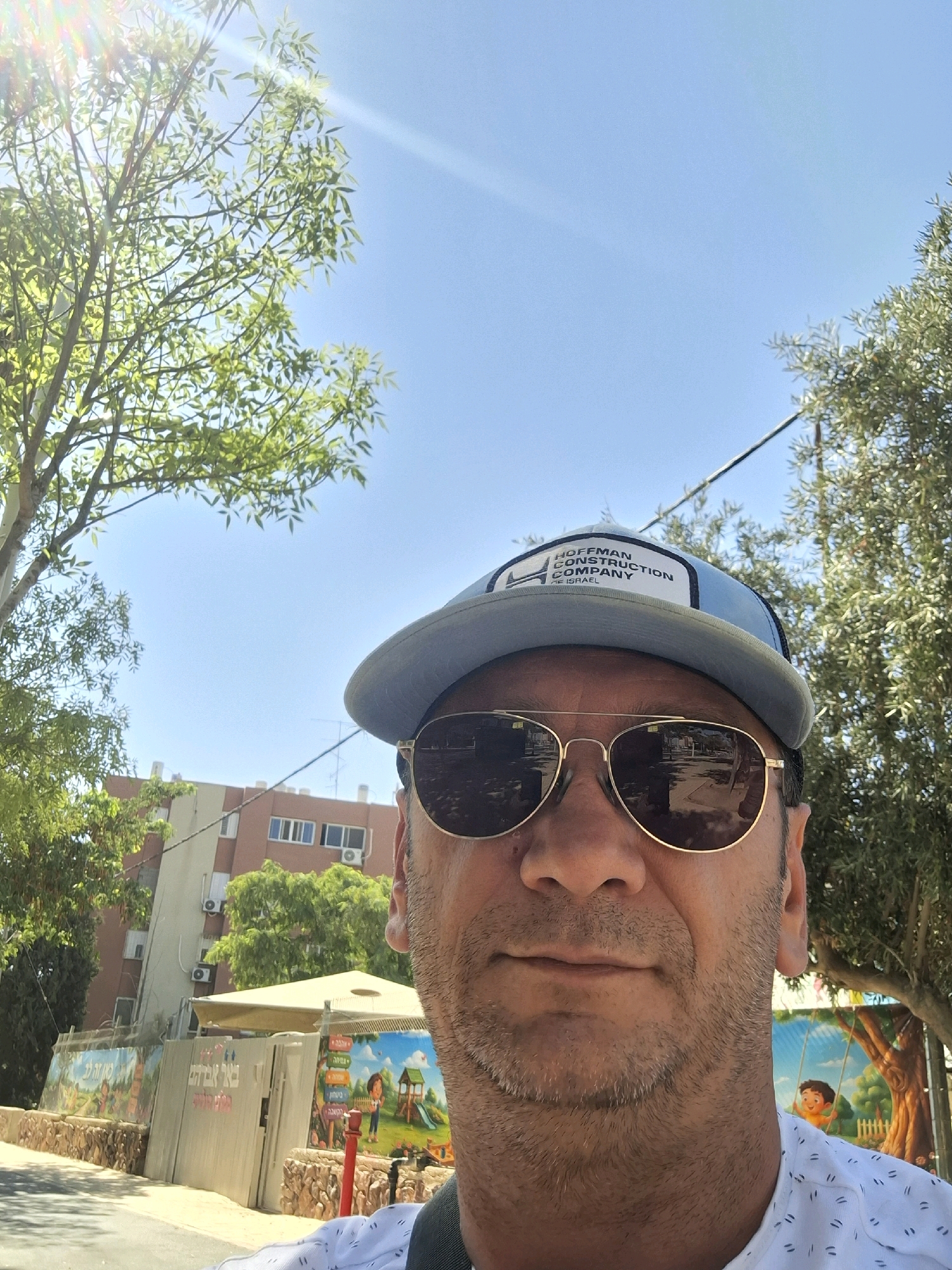 A man wearing a light blue baseball cap with a "Hoffman Construction Company" logo and aviator sunglasses stands in front of a building in Yerocham, Israel. The man is looking directly at the camera. Behind him, a tree with green leaves partially obscures a light brown and white building. In the foreground, to the left of the man, is a wall with a mural depicting children playing. The mural has text in Hebrew. The sun is shining brightly, casting a lens flare across the upper left portion of the image.