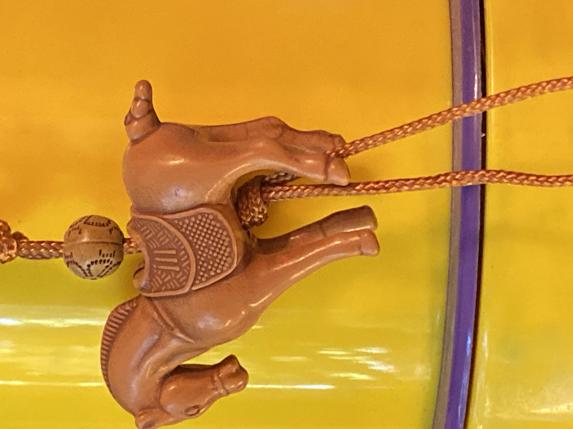 A close-up, eye-level shot captures a decorative carved wooden horse ornament hanging against a bright yellow background. The horse is a light brown color and is detailed with a saddle and reins. A small, carved, patterned bead is suspended above the horse. Below the horse, two braided cords descend. The ornament appears to be attached to or displayed on a cylindrical object with a thin purple band visible near the bottom of the frame. The lighting is bright, creating some glare on the yellow surface. There are no people or any identifying features of Ivanteyevka, Russia, in this image.