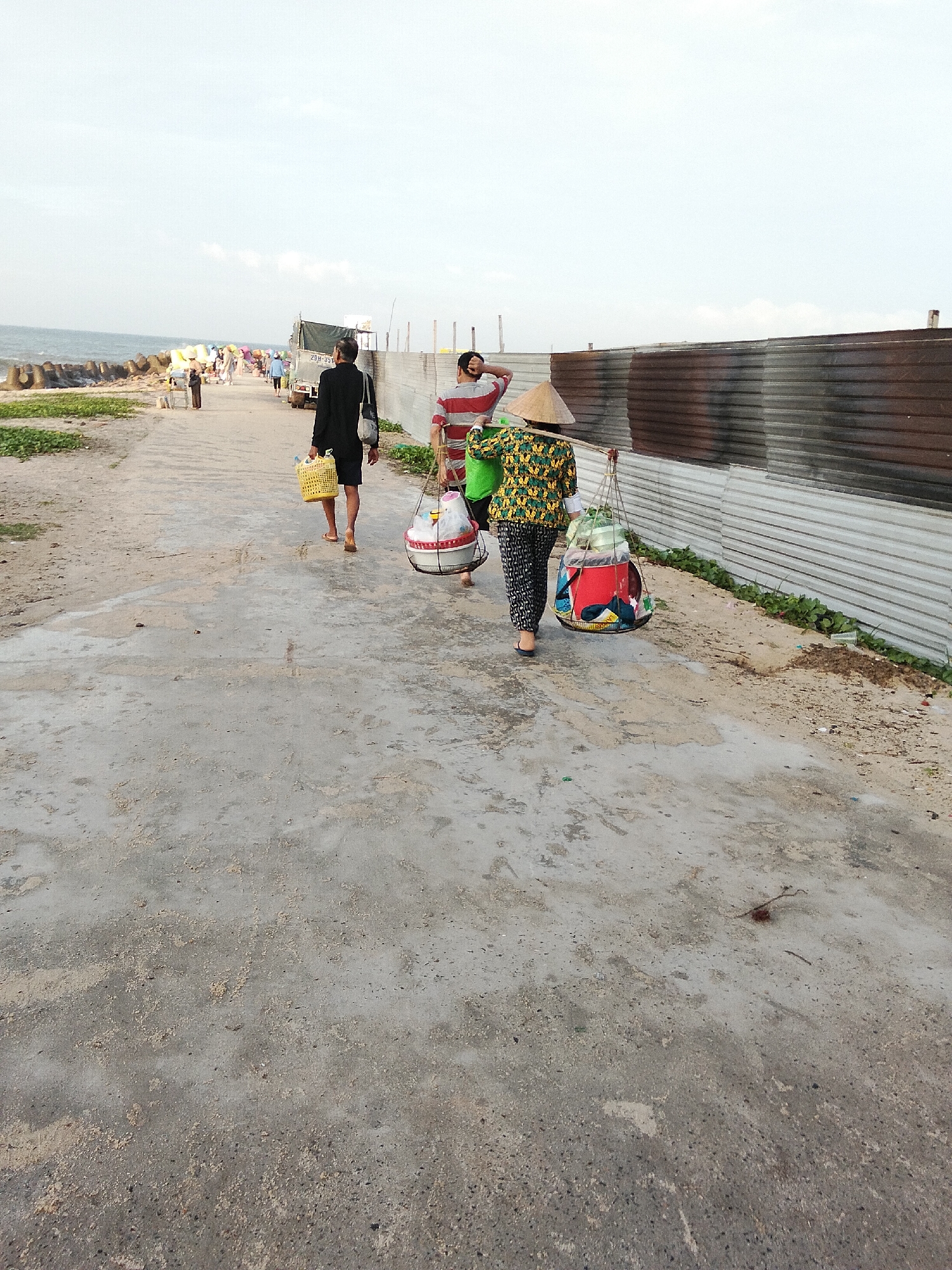 The image captures a scene along a coastal path in Phú Thủy, Vietnam. The path is made of concrete and appears weathered, with scattered sand and debris. To the left, the ocean is visible with a breakwater made of large concrete tetrapods, and beyond that, a collection of small stalls and people suggest a market or gathering. In the background, a corrugated metal fence runs along the right side of the path, separating it from an area with some vegetation.
In the foreground and mid-ground, three individuals are walking away from the camera. The person closest to the camera, on the right, is a woman wearing a conical hat, a patterned long-sleeved shirt, and patterned pants, carrying a large basket suspended from a pole across her shoulders. The basket contains various items, including a red container and what appear to be fabrics or bags. To her left, a second person, possibly male, is walking in the same direction, carrying a yellow woven basket in their right hand. Further ahead, a third person, also possibly male, is walking away, dressed in a dark outfit and holding a yellow basket. Other people and stalls are visible further down the path, indicating a populated area. The sky is overcast.