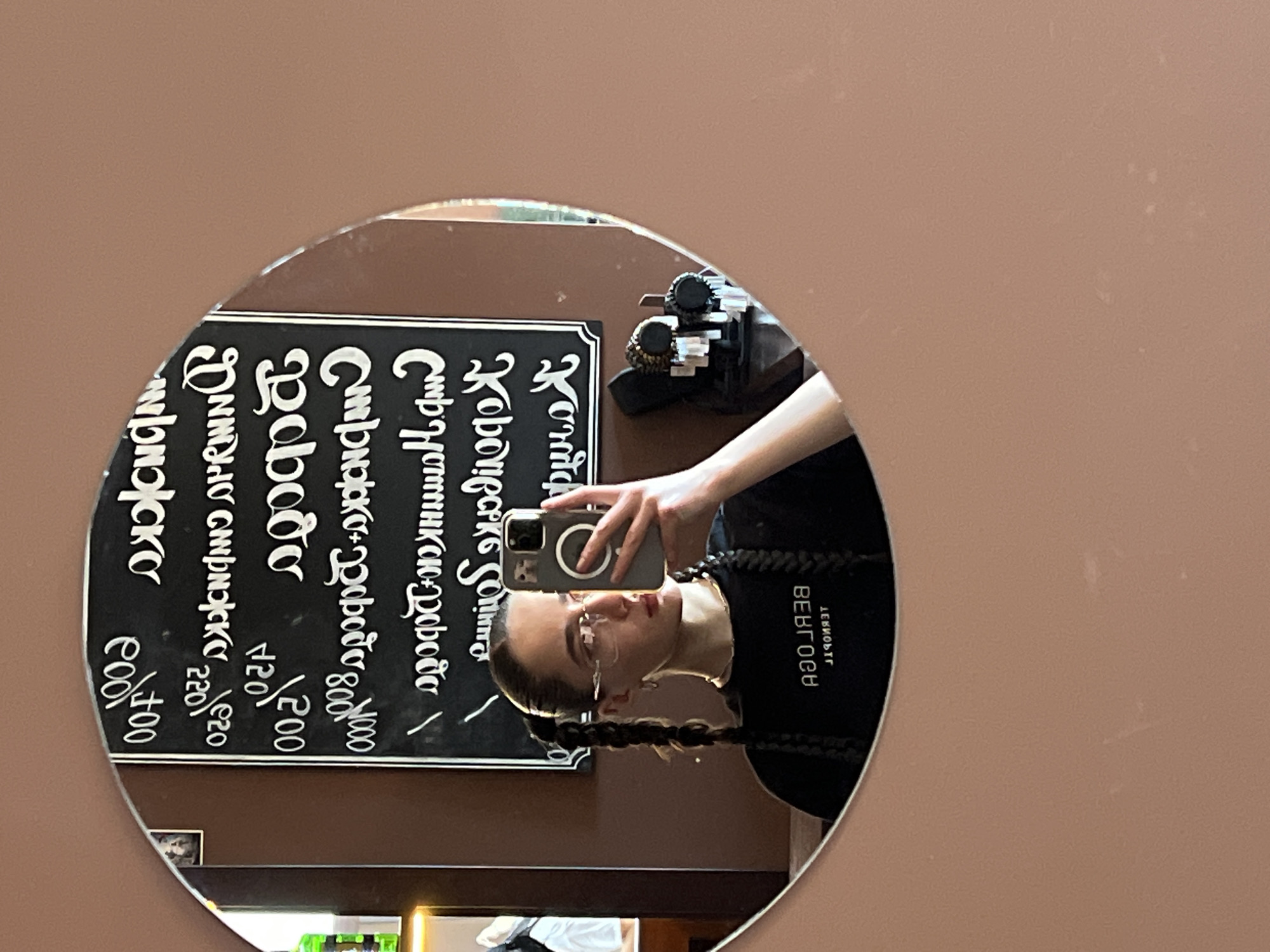 A young woman with dark hair styled in two braids and wearing glasses is reflected in a round mirror. She is holding a smartphone up to take the picture. She is wearing a black t-shirt with white Cyrillic text on the back that reads "ДЕВИОЬТГ ВЕВГОСУ". In the mirror's reflection, behind her, is a blackboard with white handwritten text in Cyrillic. The text appears to be a menu with prices, featuring items such as "Стрижка-ревода 80000", "Poboro 2200", and "рижка Coditoo 22020". There are also some prices listed next to items in what appears to be Japanese Yen, such as "100円". The setting appears to be a salon or barbershop, suggested by the menu and the hairstyle services mentioned. The overall lighting suggests it is daytime. There are no strong emotional cues visible, but the woman seems relaxed. The location context of Ternopil, Ukraine is provided, which aligns with the Cyrillic script on the menu.
