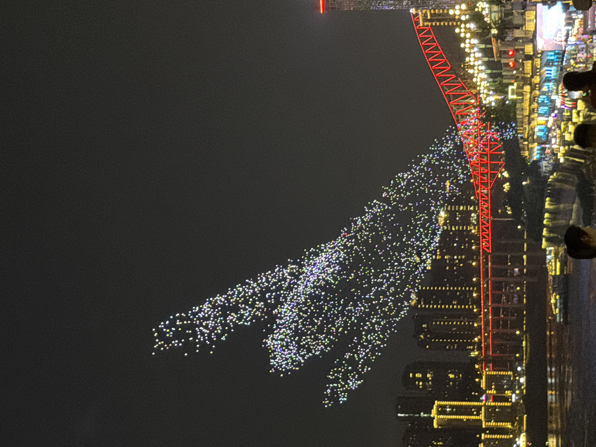 This image captures a nighttime scene along the Yangtze River in Nan'an District, Chongqing, China. The sky is illuminated by a drone light show that has formed the shape of a hand. The city skyline features numerous high-rise buildings with illuminated windows and external lighting in yellow and white. A prominent red bridge spans the river, adorned with festive lights. The foreground shows a body of water, likely the Yangtze River, with a few small boats visible. On the riverbank, there are more buildings and lights, creating a vibrant urban landscape. Two individuals, partially visible in the lower right corner, are facing away from the camera, observing the spectacle. The overall atmosphere is one of celebration and visual spectacle.