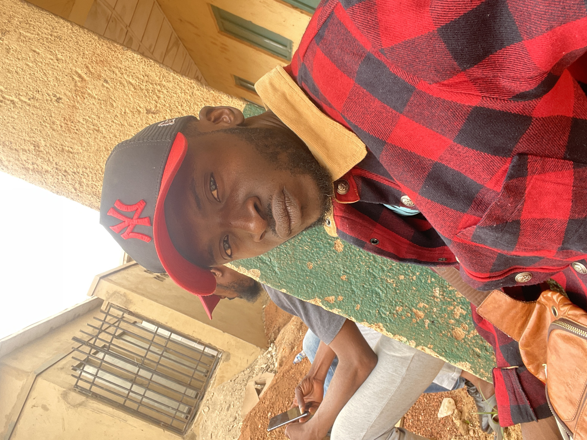 The image shows a man in a red and black flannel shirt and a black baseball cap with a red brim and a white "NY" logo. He is leaning on a green concrete ledge. Another man sits in the background, using a mobile phone. The environment includes a building with barred windows and an earthen ground. Based on the visual context, it is plausible that this image was taken in Jos, Nigeria.