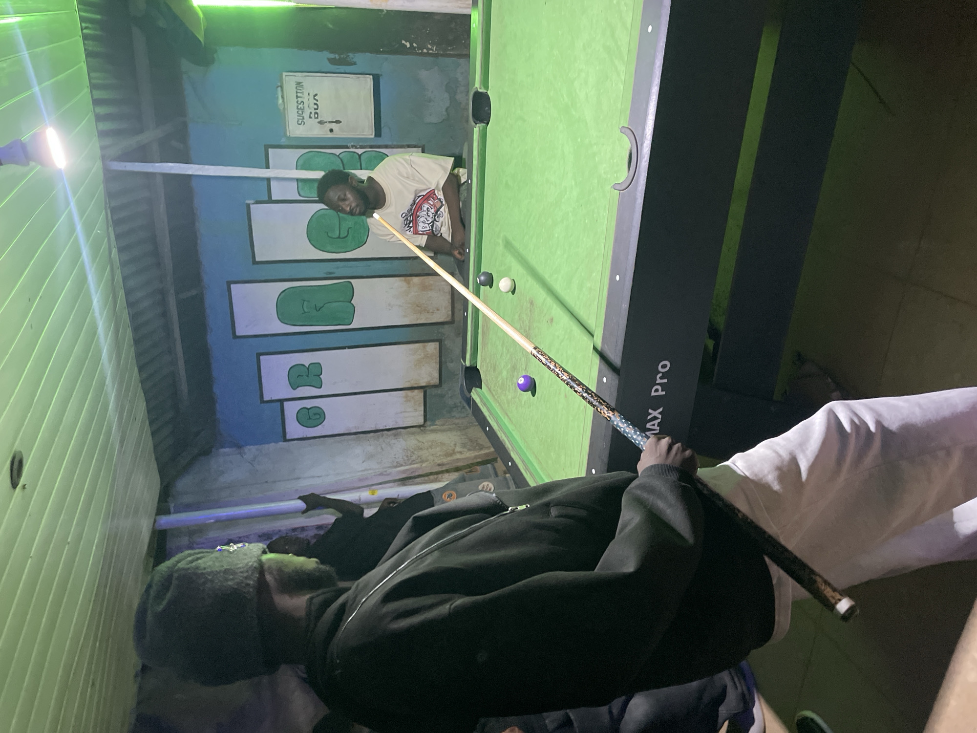 A medium, indoors shot reveals two individuals playing pool inside what appears to be a bar or pool hall in Durimi, Nigeria. One person, wearing a light t-shirt, is poised to strike a cue ball on a green pool table towards an 8-ball. The other person, wearing a dark hooded sweatshirt and a dark cap, is holding the cue stick. The setting includes posters or painted artwork on the wall behind the player and overhead lighting. The pool table has the words "MAX Pro" on the edge. The hall's architecture features corrugated metal roofing, likely as a sound and lighting treatment.