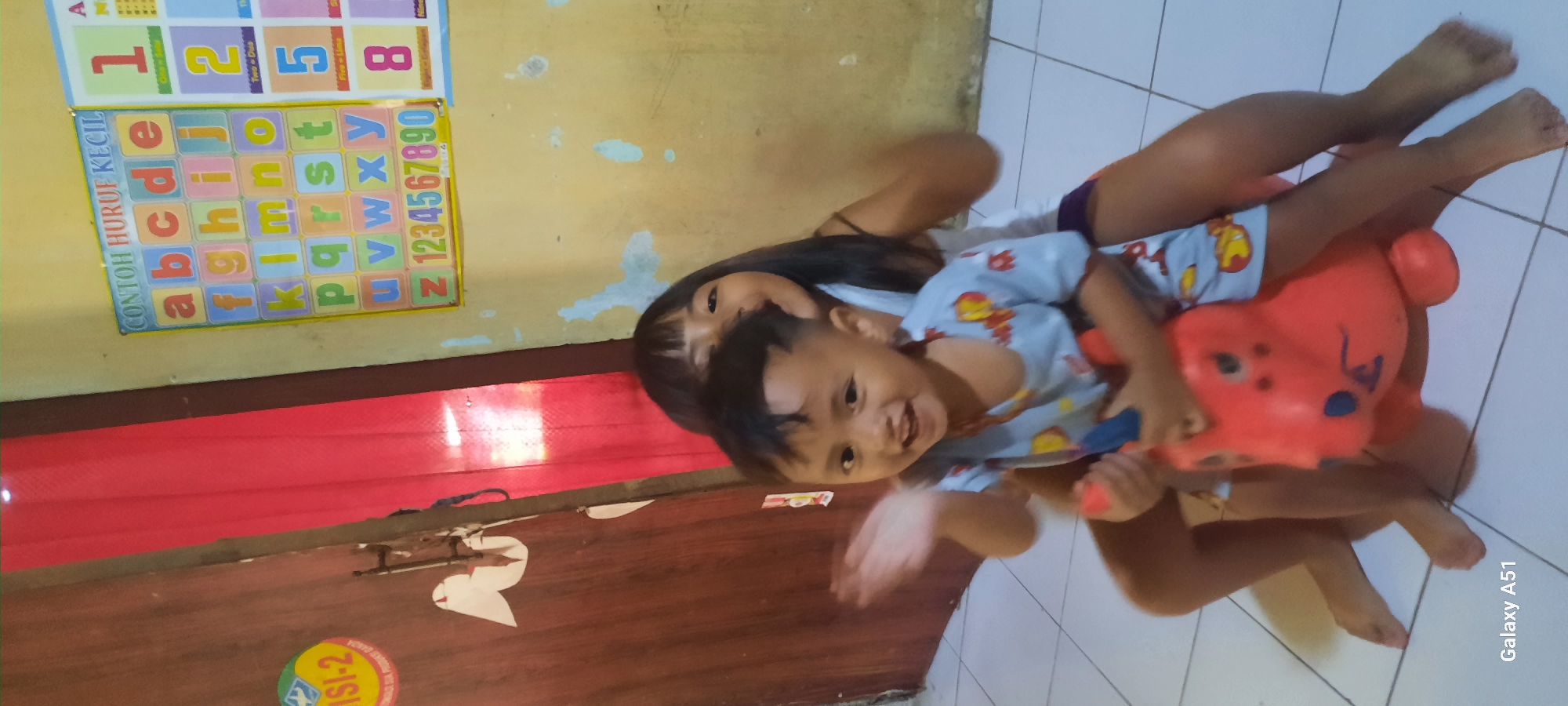 In Ploso, Indonesia, a young boy is seated on a red toy animal, possibly a rocking horse, with his hand raised in a wave. He is wearing a light blue pajama set with a cartoon pattern. Behind him, a young girl is partially visible, her face smiling and her dark hair framing her face. They appear to be in a room with a light-colored tiled floor. To the left, a portion of a wooden door is visible, adorned with white cutouts resembling birds. On the wall to the right, educational posters featuring the alphabet and numbers are displayed. The lighting suggests an indoor environment, likely a home.