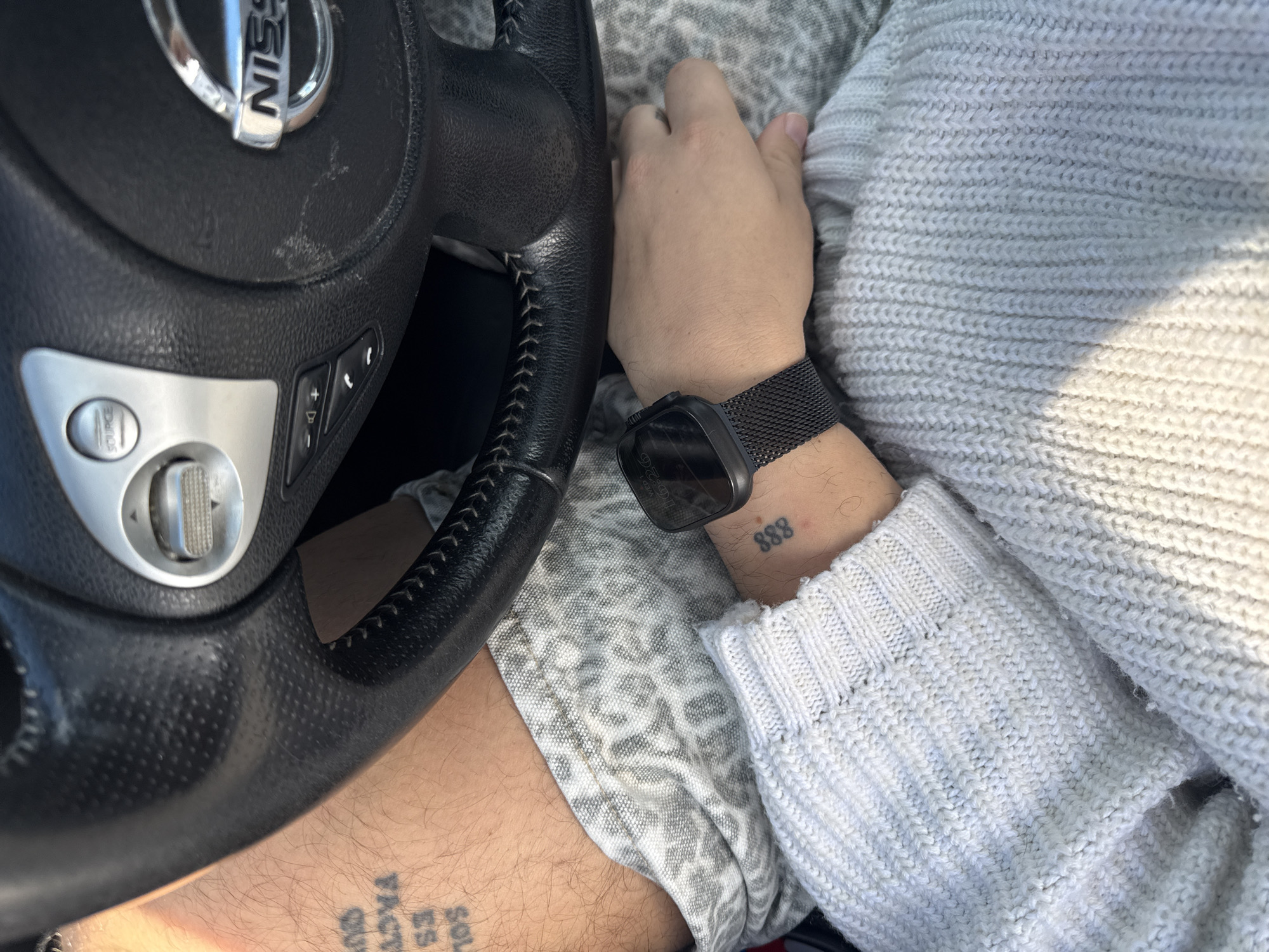 A person is sitting in the driver's seat of a Nissan vehicle. Their left hand is on the steering wheel, while their right hand rests on their lap. The person is wearing a gray sweater and a black smartwatch. Tattoos are visible on both forearms. The location is not specified in the image, therefore it is not known if the location is in the city of Esch-sur-Alzette, Luxembourg.