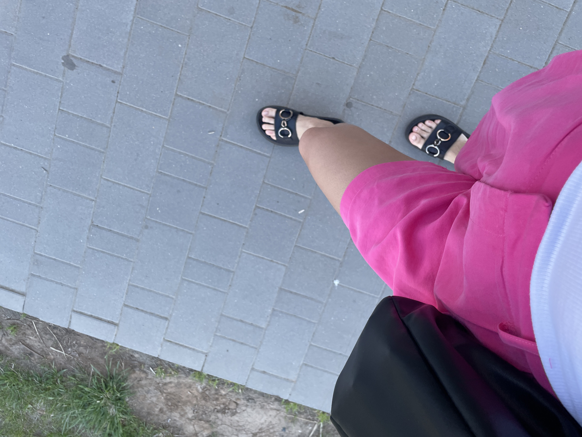 A top-down shot shows a person standing on a gray tiled sidewalk. The person is wearing a pair of black sandals with gold circular embellishments. They are also wearing bright pink shorts and a white shirt. A black bag is visible beside the person's leg. The edge of the sidewalk is next to a grassy area. The photo does not show enough information to determine the location is Izhevsk, Russia.