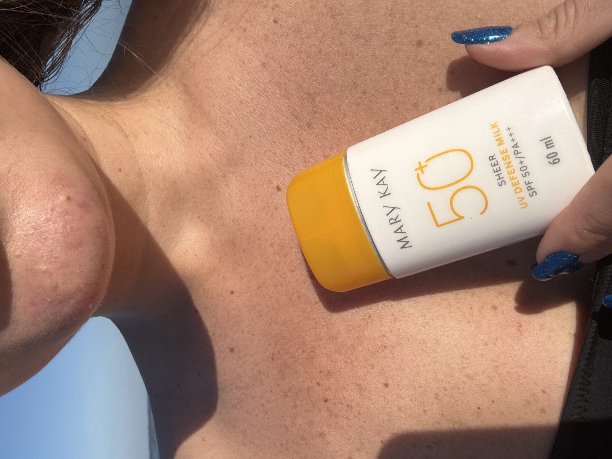 The image shows a close-up view of a person's chest and neck, with a bottle of Mary Kay sunscreen. The person has fair skin with freckles and is wearing a black swimsuit. The bottle is white with an orange cap and displays the text "MARY KAY", "50+", "SHEER UV DEFENSE MILK SPF 50+/PA+++", and "60 ml". The background is a blurred blue sky, suggesting an outdoor setting. The location of Denpasar, Indonesia is not depicted in the image.